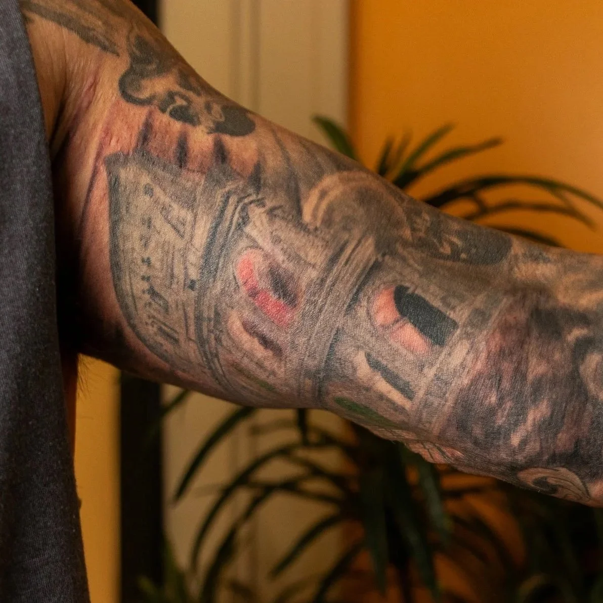 Close-up of a tattooed arm featuring various black and red ink designs, with a background of indoor plants.