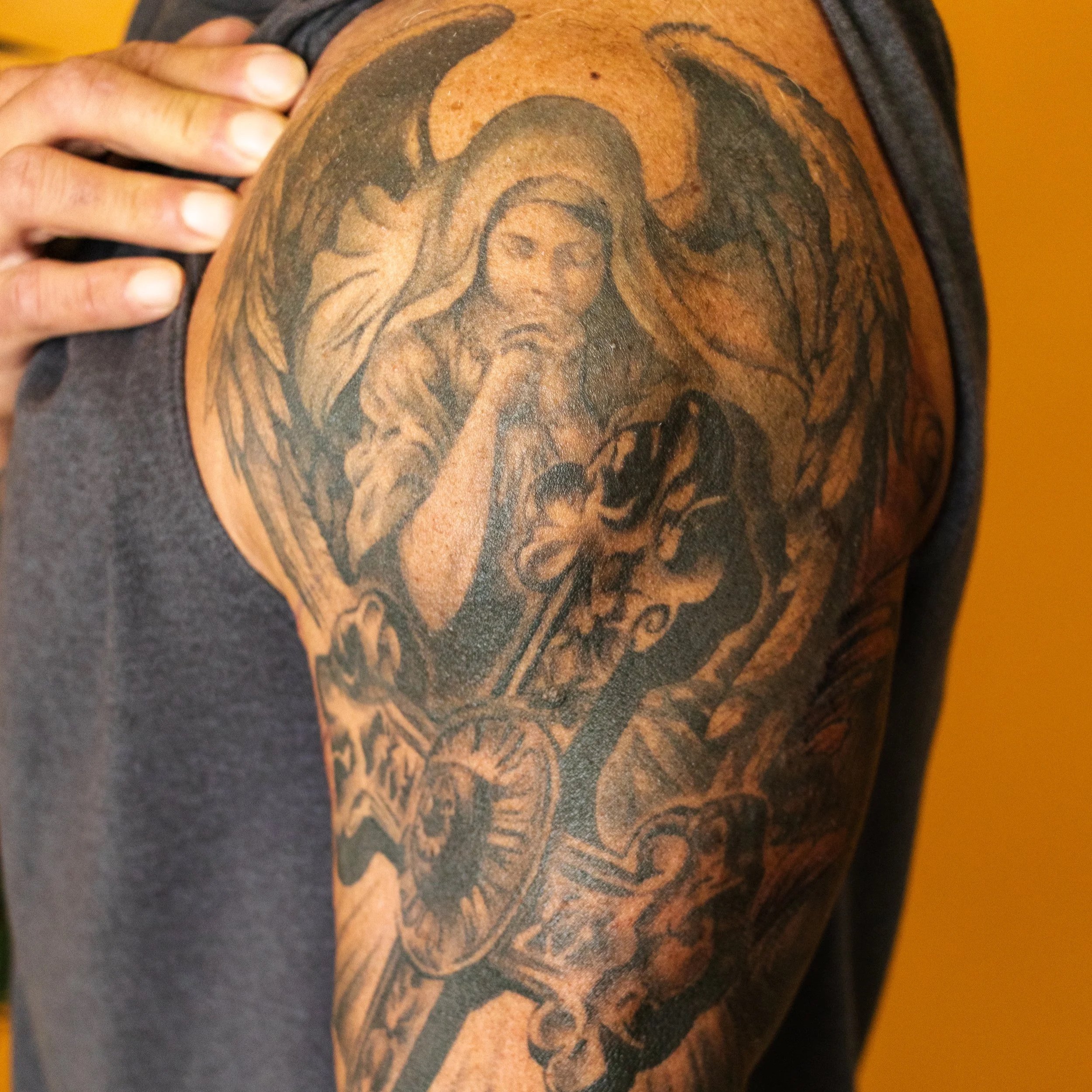 A tattoo on a person's upper arm depicting a woman with long hair, holding a sword and surrounded by angel wings, a clock, and a skull.