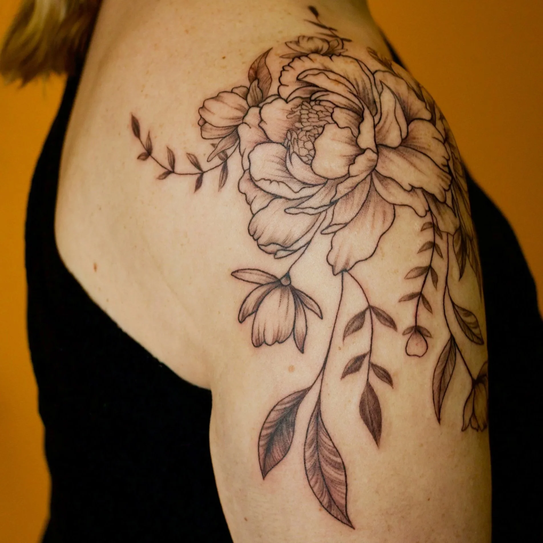 Black and gray floral tattoo of a large peony flower with leaves and smaller flowers on upper arm near shoulder.