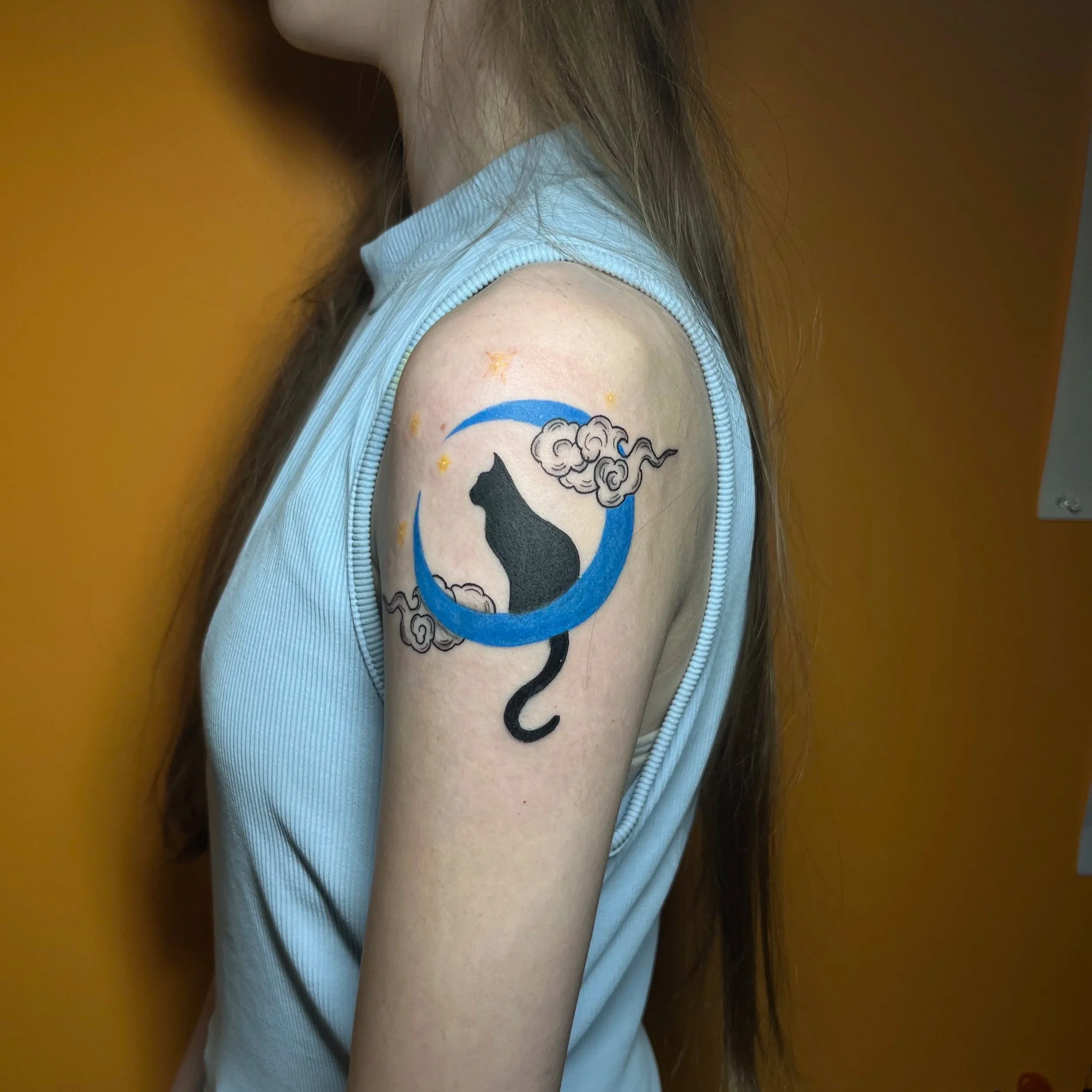 Tattoo of a black cat sitting on a blue crescent moon with clouds and stars on a person's upper arm.