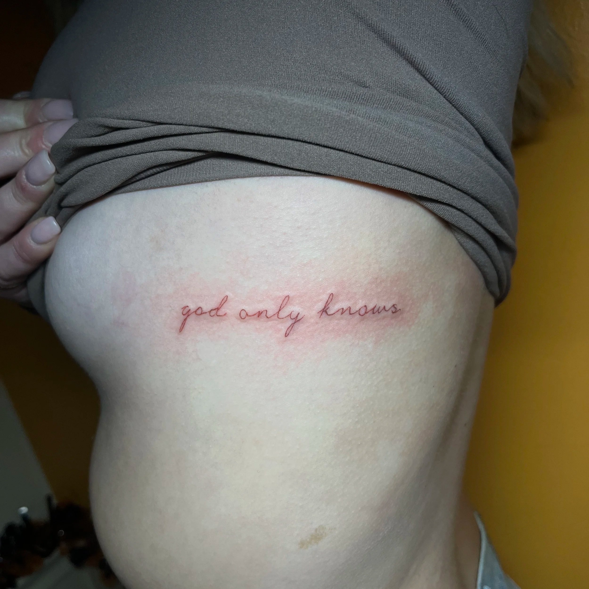 A person's side upper body with a tattoo that reads 'god only knows' in red cursive ink, located on the side of the torso near the ribcage.