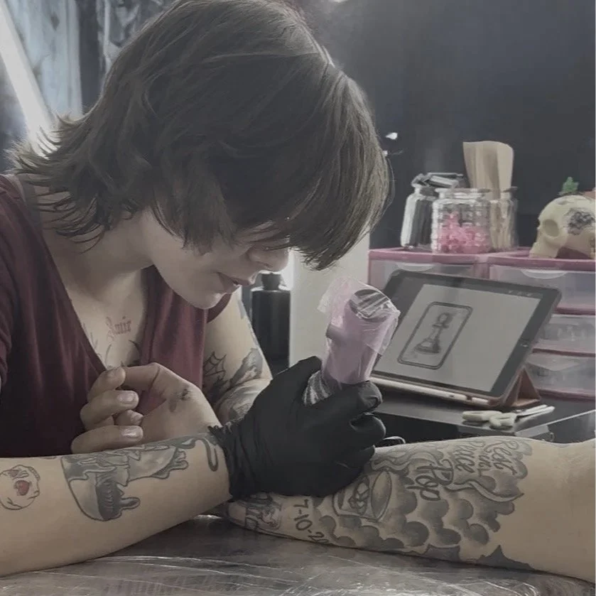 Tattoo artist working on client's arm, holding tattoo machine, in a tattoo studio with equipment and artwork in the background.