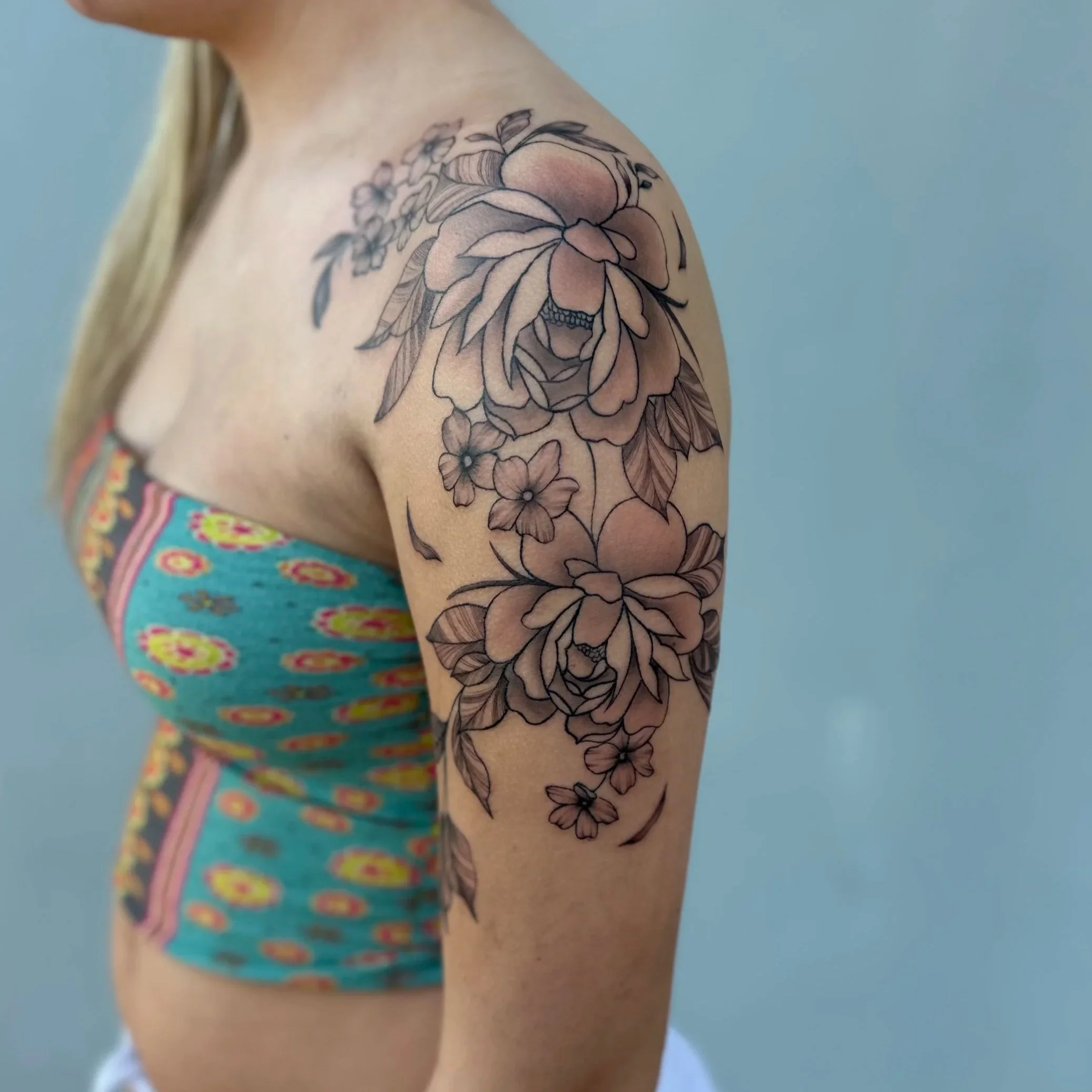 Close-up of a woman's upper arm and shoulder showing a large floral tattoo on her upper arm. The tattoo features detailed roses, smaller flowers, and leaves in black and gray ink.
