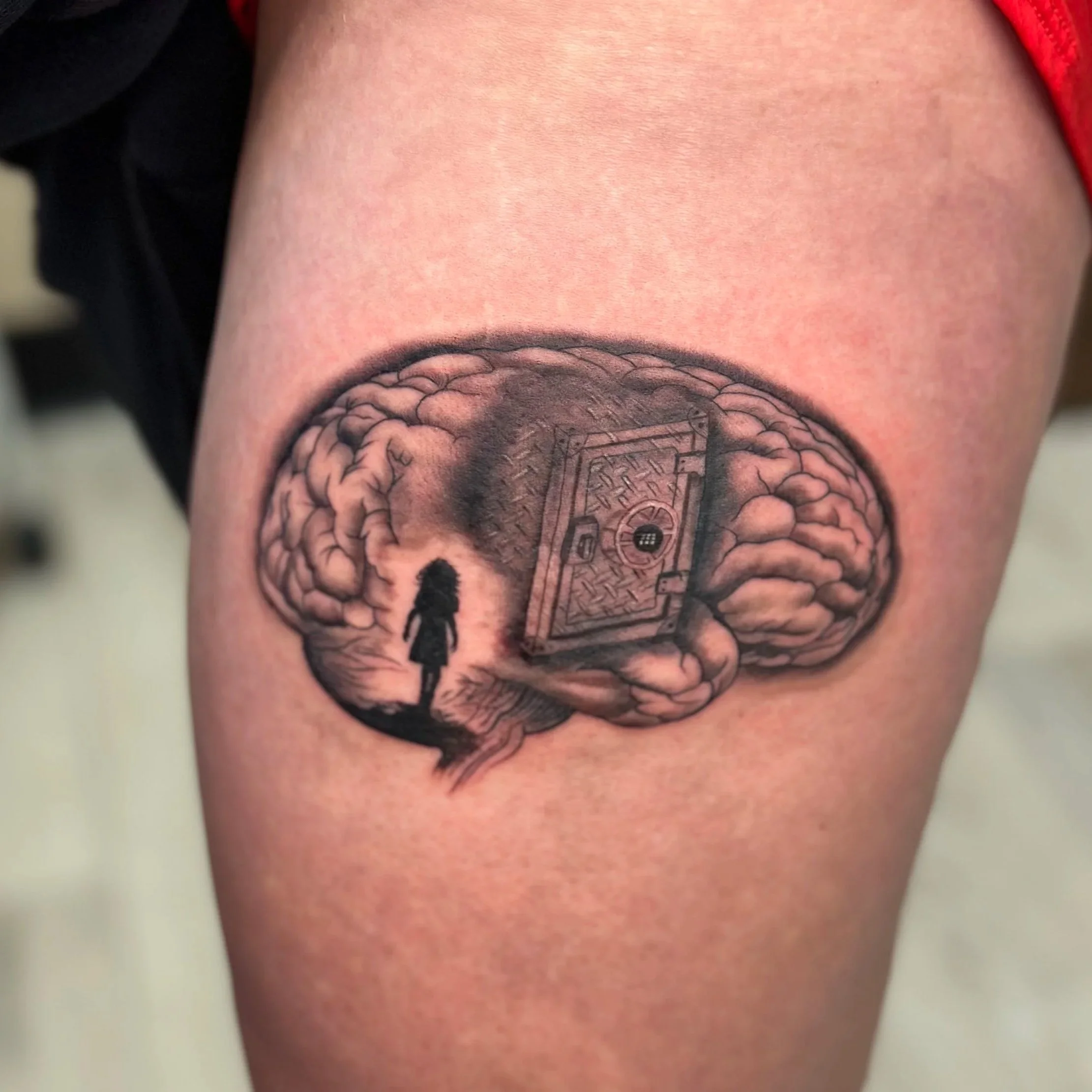 Tattoo of a brain with a safe inside it, featuring a silhouette of a girl walking towards it, created with black and gray ink.