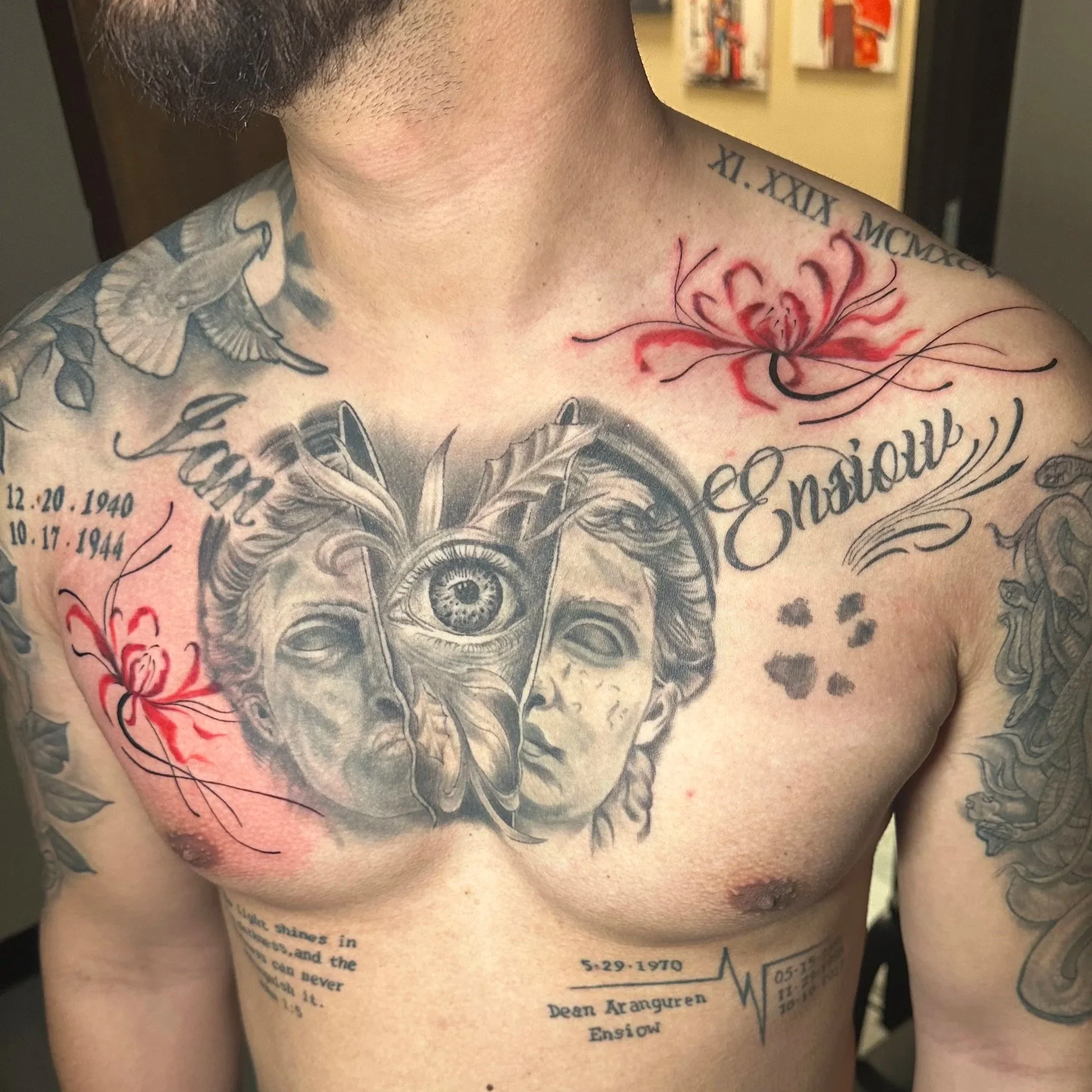 A man with multiple tattoos on his chest and shoulders, including a large black and gray tattoo of a woman's face with an eye in the center, red flowers, and script text.