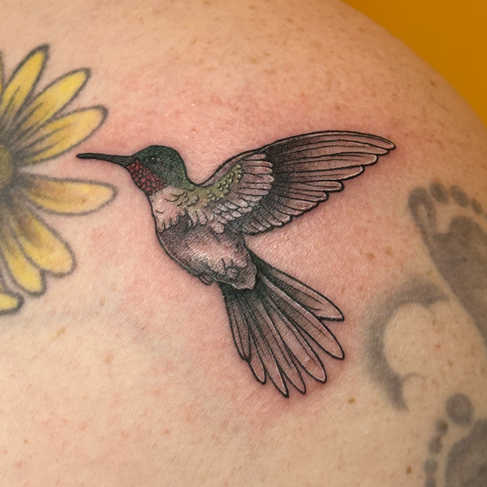 Colorful tattoo of a hummingbird hovering near yellow flowers on a person's skin.