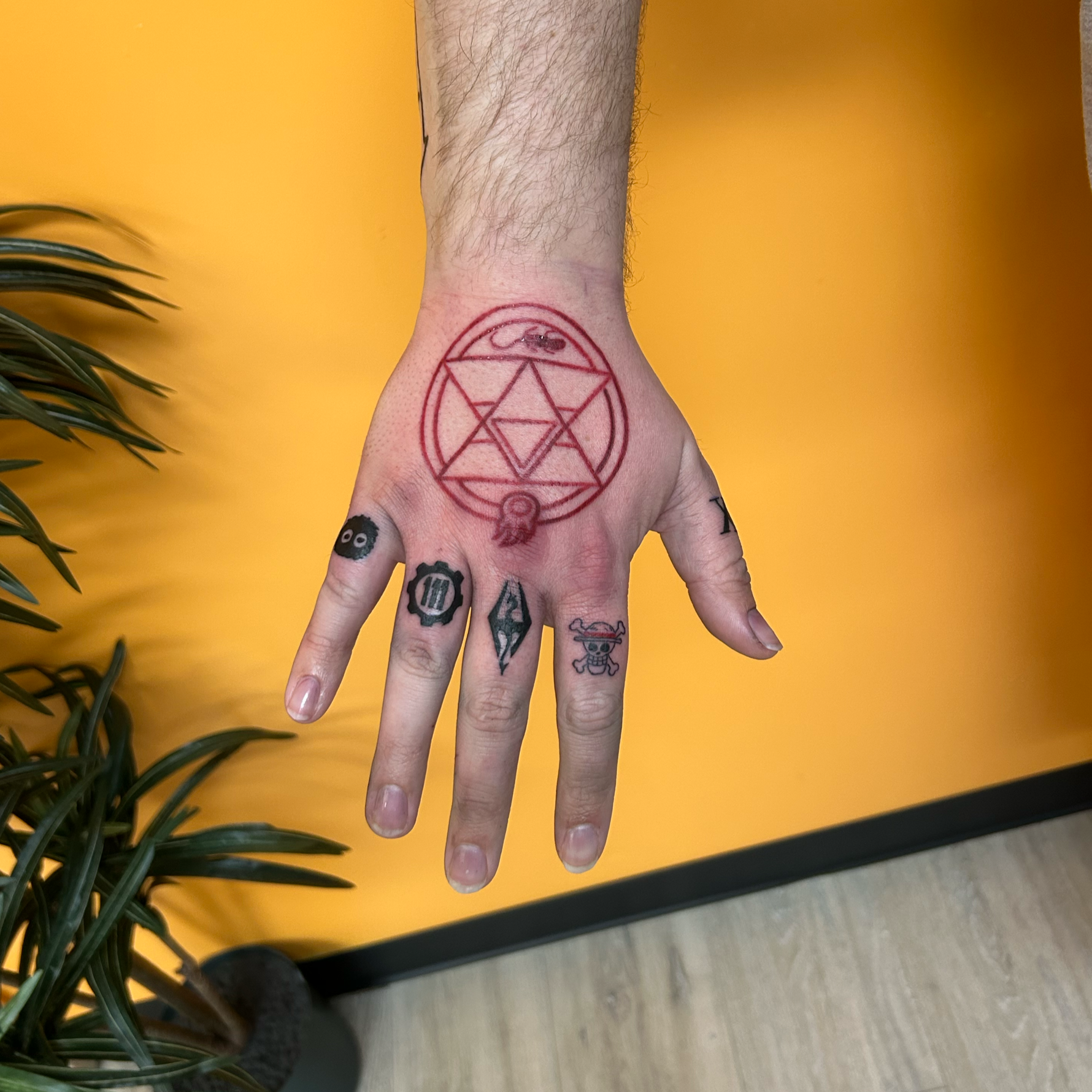 A person's hand with various tattoos, including a large red geometric design on the wrist, set against an orange background with some green plant leaves visible on the left.