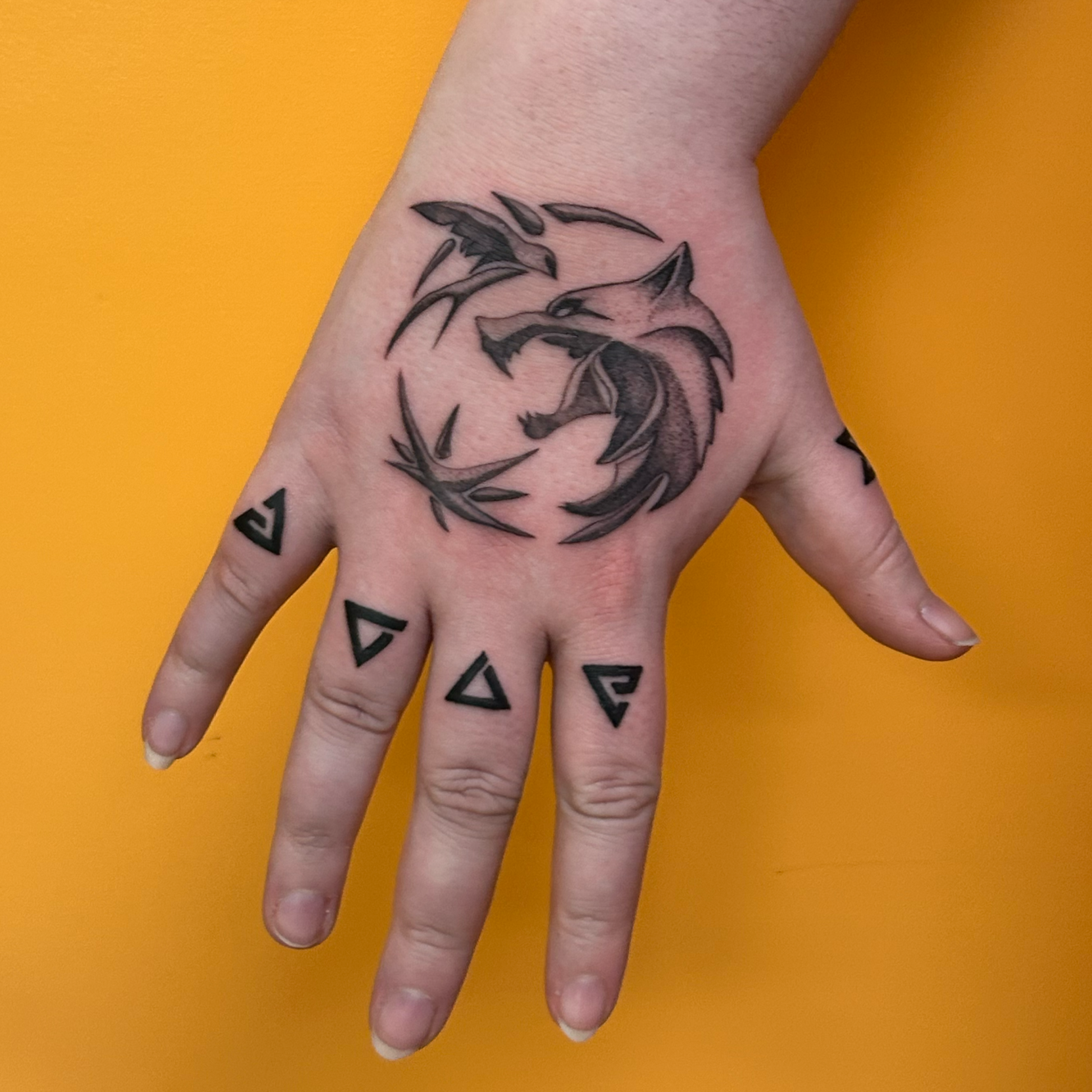A hand with several tattoos, including a dragon tattoo on the back of the hand and geometric shapes on the fingers, against a yellow background.