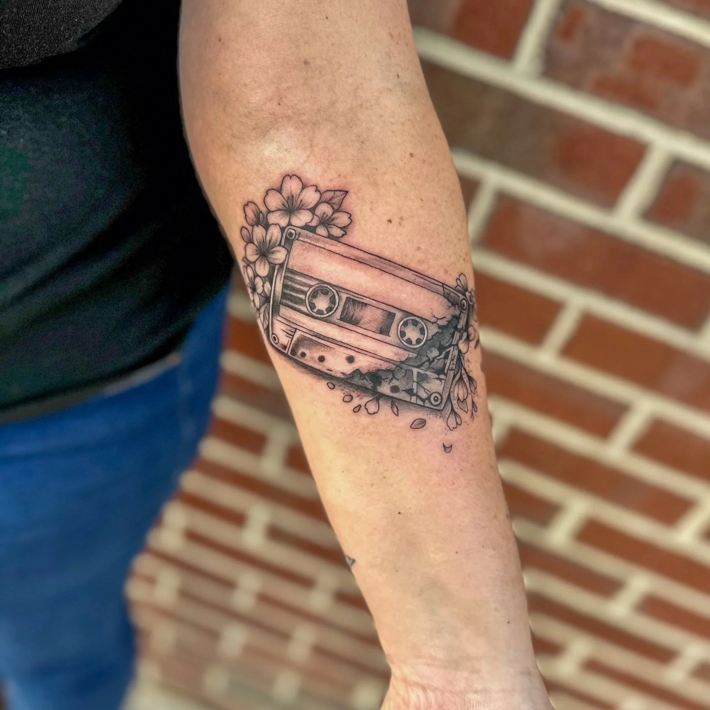 Close-up of a person's arm with a tattoo of a flower and a cassette tape, with a blurred brick wall background.