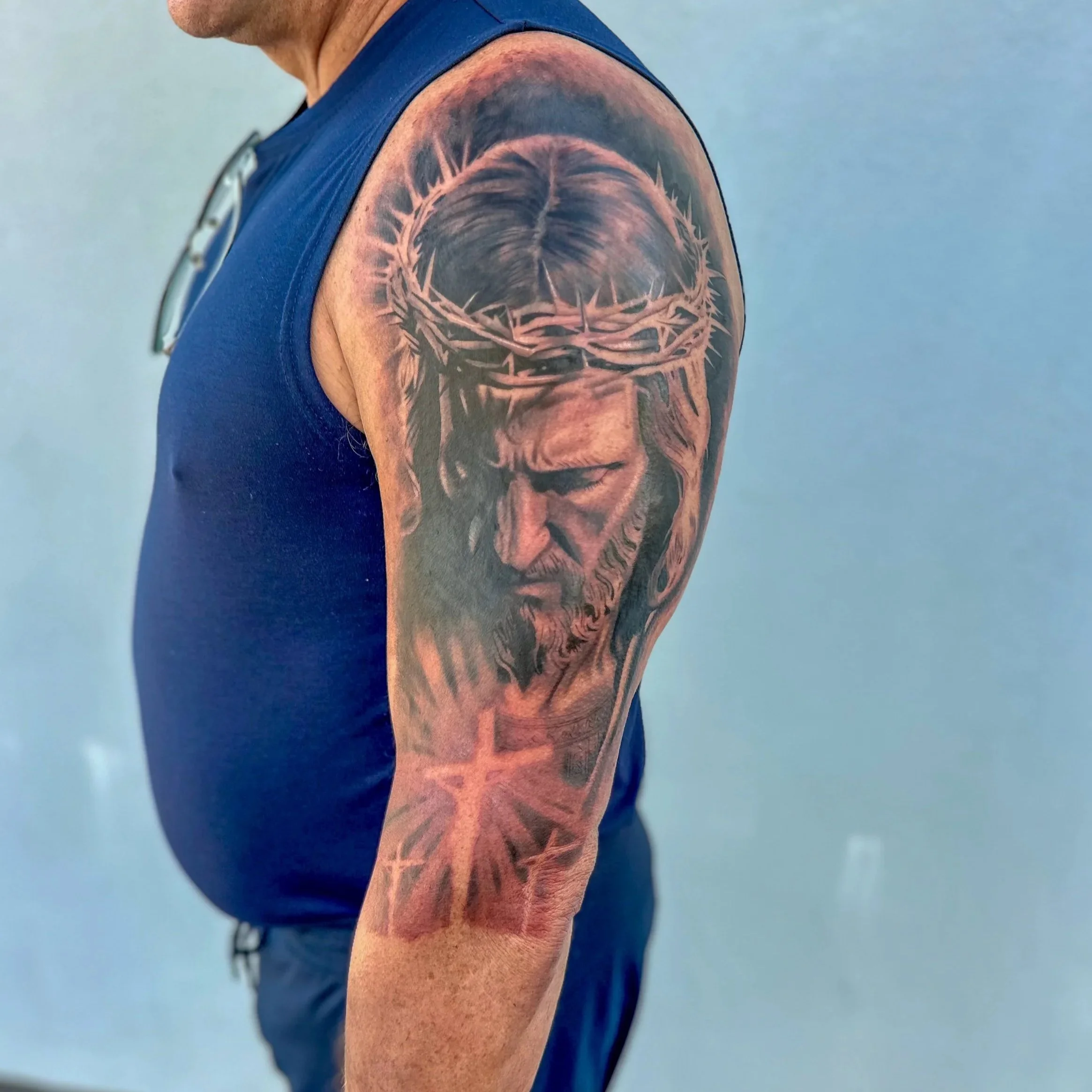 Close-up of a person's upper arm and shoulder with a detailed tattoo of a crown and clenched fists, wearing a blue sleeveless shirt.