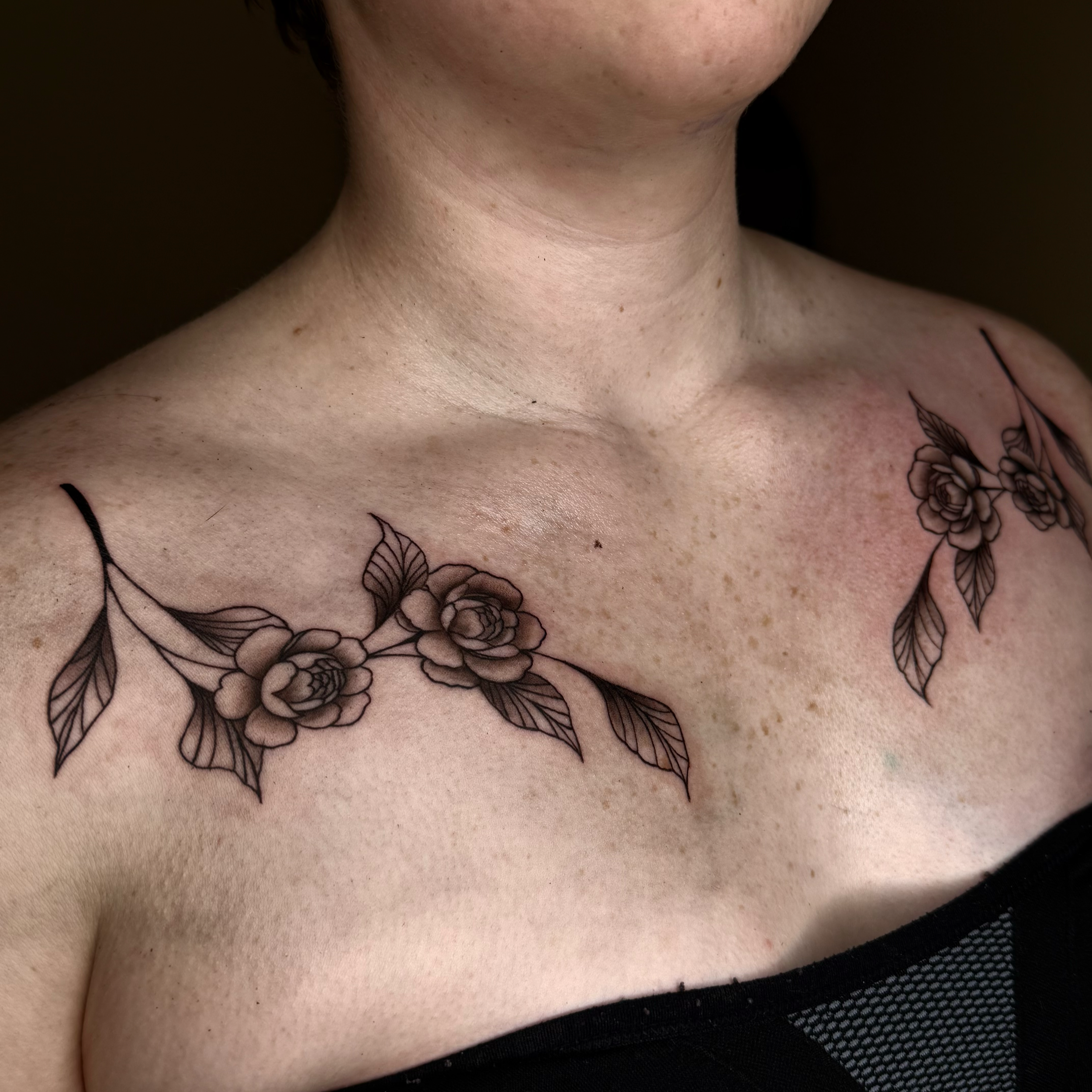 Close-up of a woman's shoulder and upper chest displaying a floral tattoo with roses and leaves on both sides of her collarbone.
