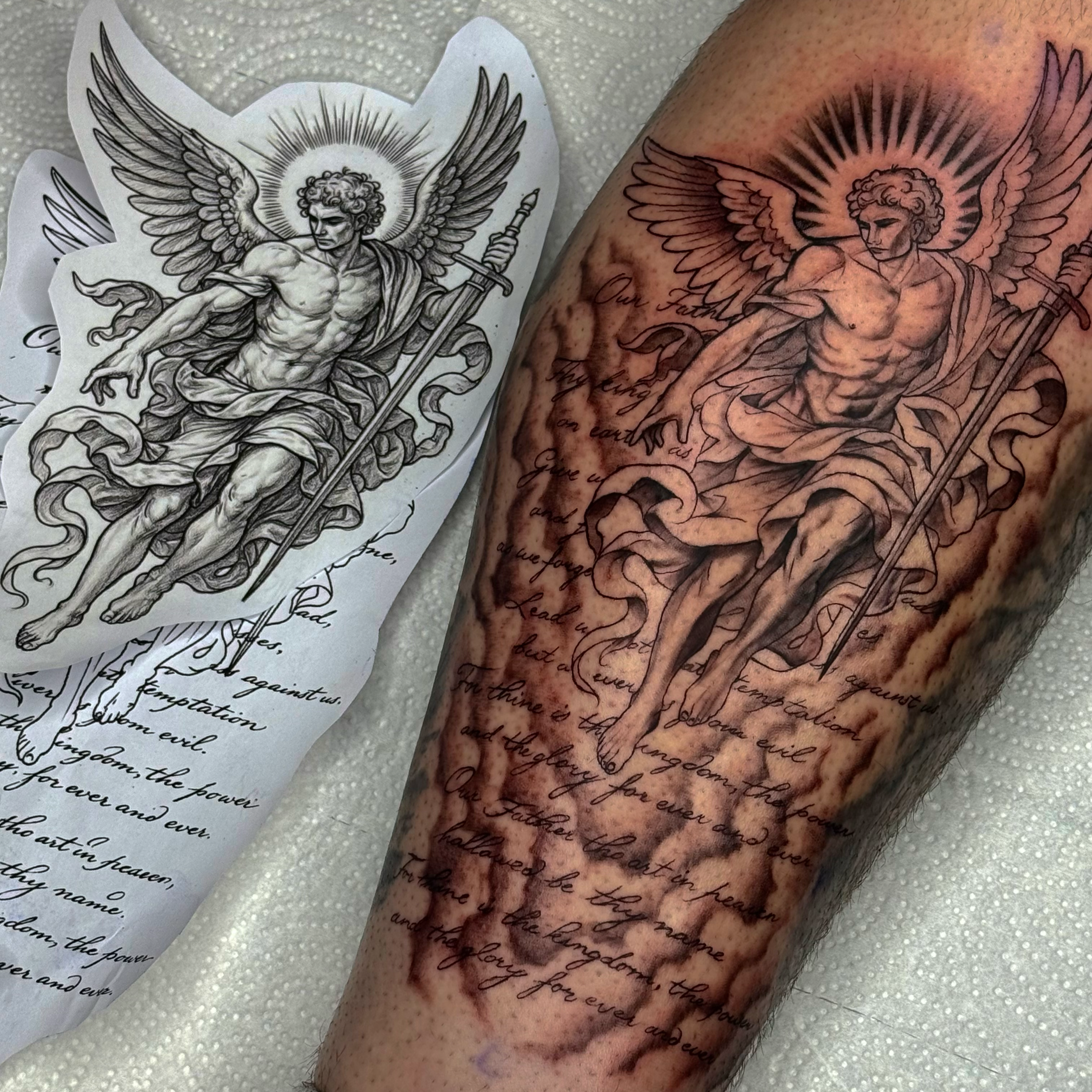 A tattoo of an angel with wings, holding a sword, with detailed shading and script writing, in progress on a person's arm.