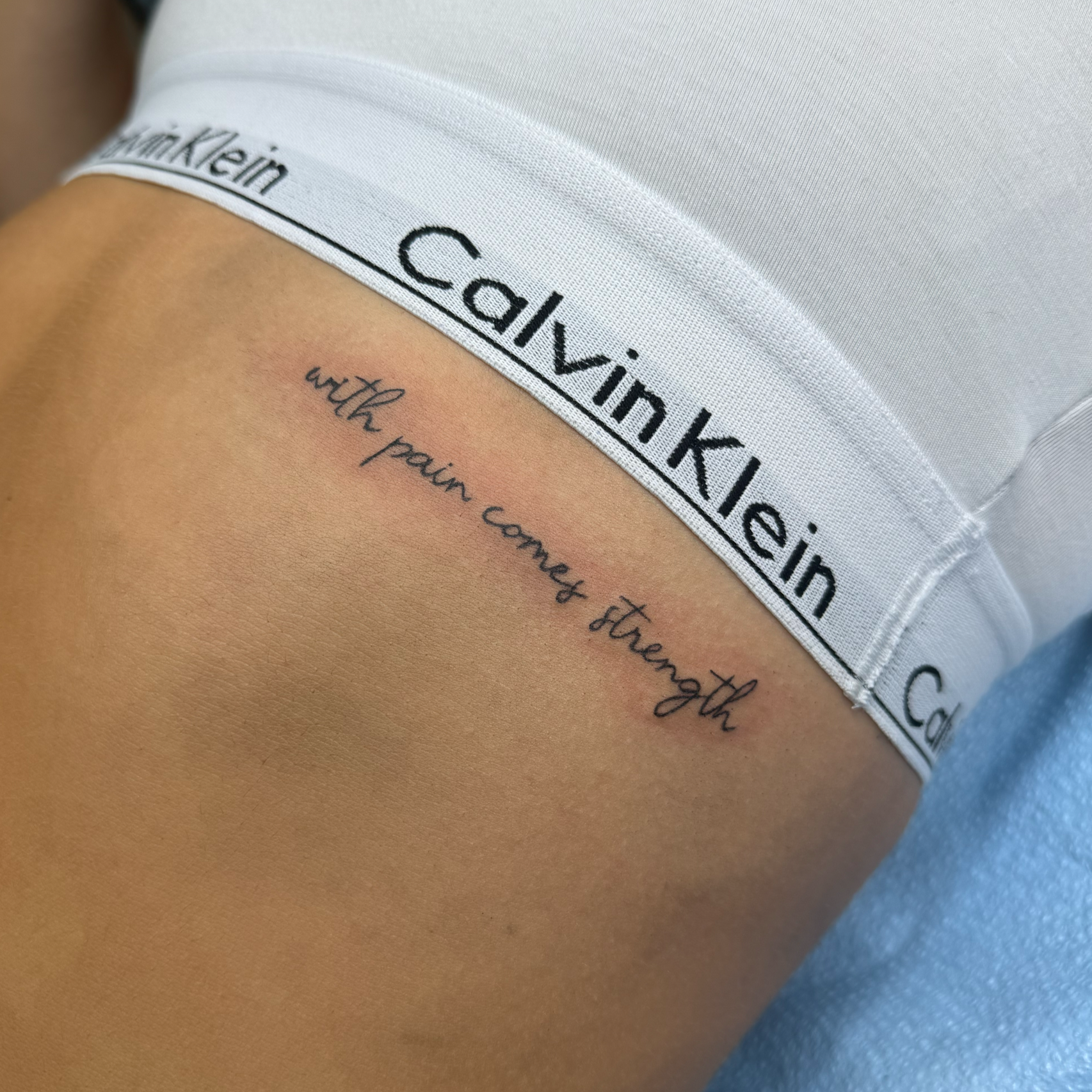 Close-up of a person's midsection wearing white Calvin Klein underwear with black text, revealing a tattoo that reads 'with pain comes strength' on the skin above the underwear