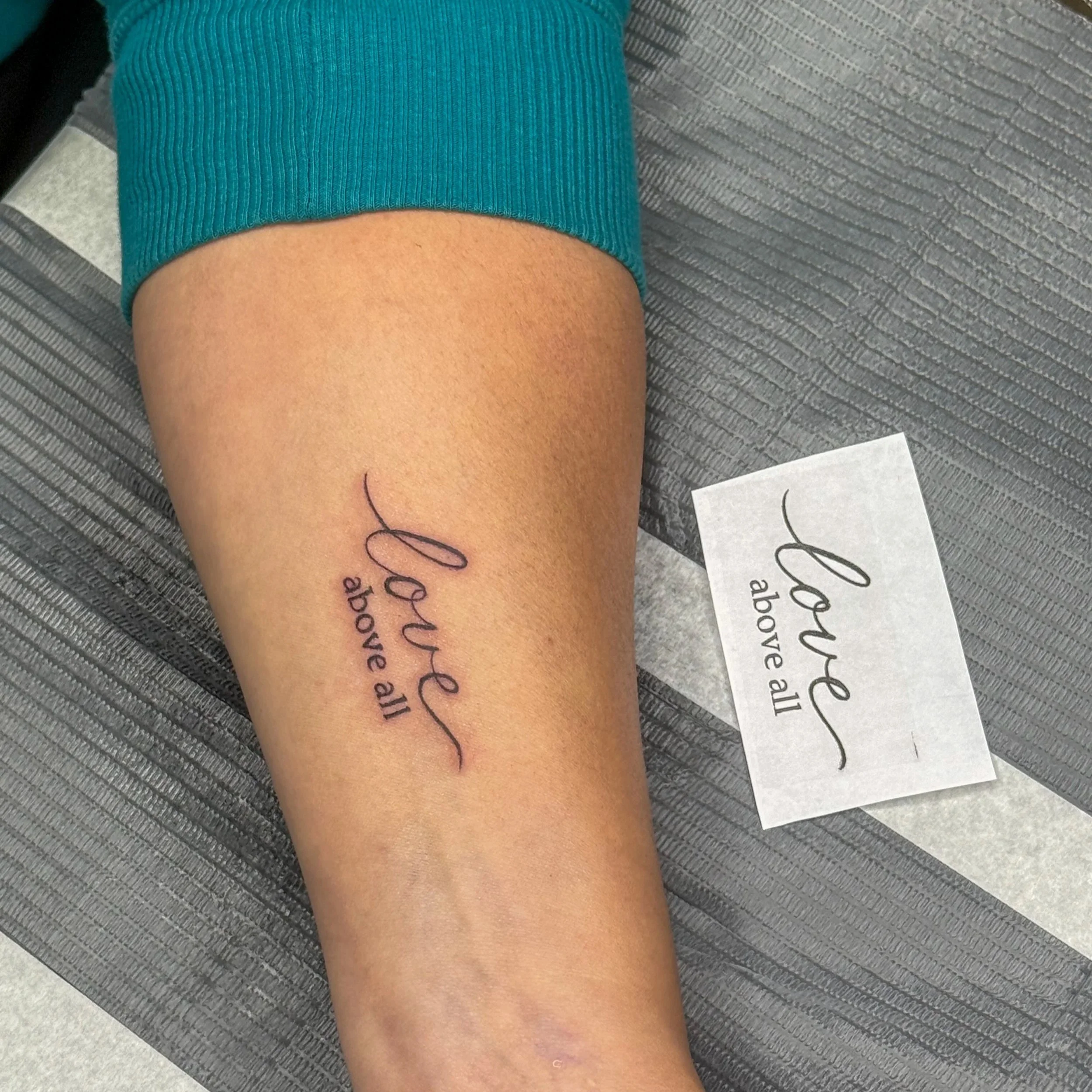 A tattoo on a person's forearm that reads, 'Love above all' in cursive script.