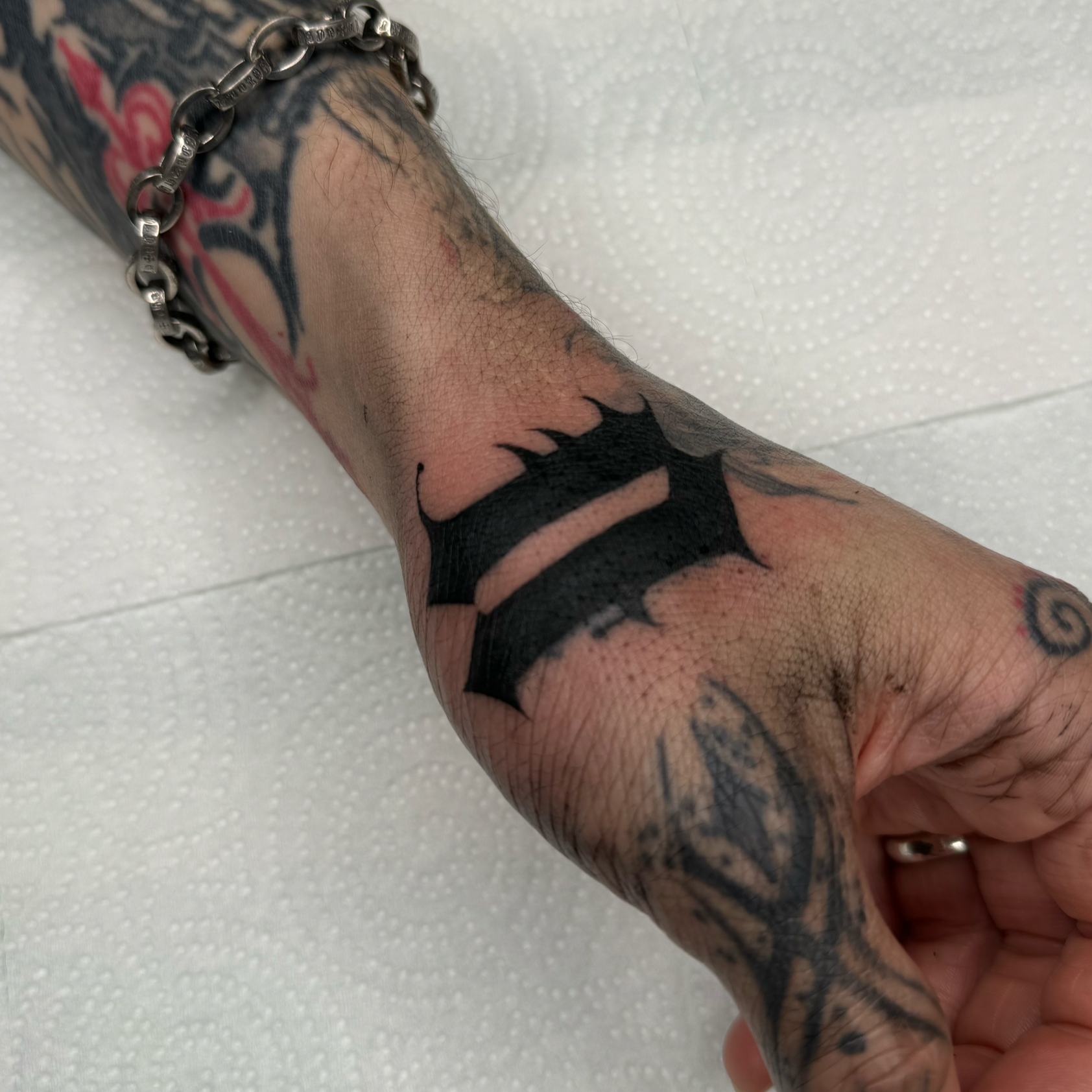 Tattooed hand with a chain bracelet resting on a white textured surface.