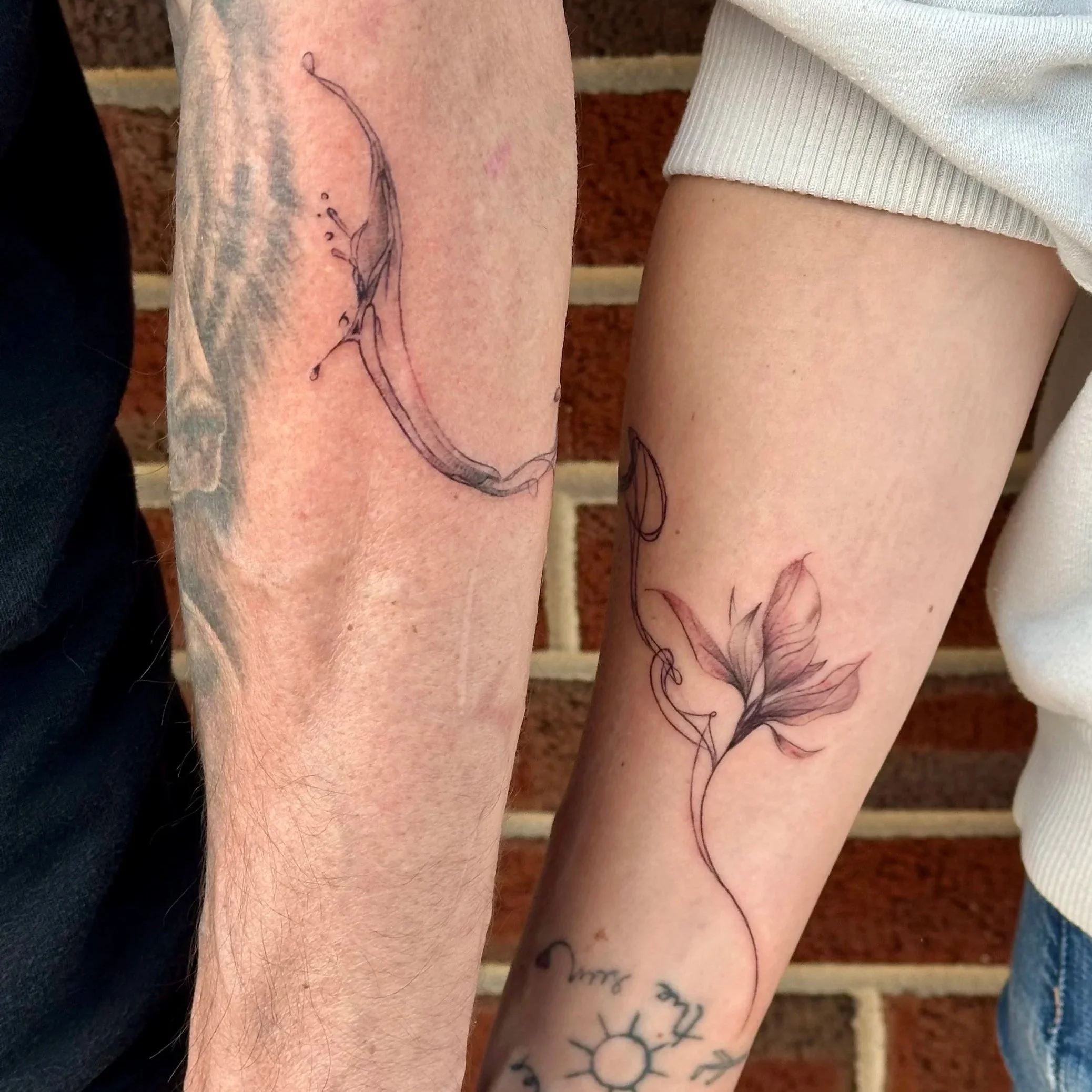 Four people’s arms with tattoos; one arm has a delicate flower tattoo, another has a sun and moon tattoo, and the other arms have various designs including a skull and abstract art; the background shows wooden stairs.