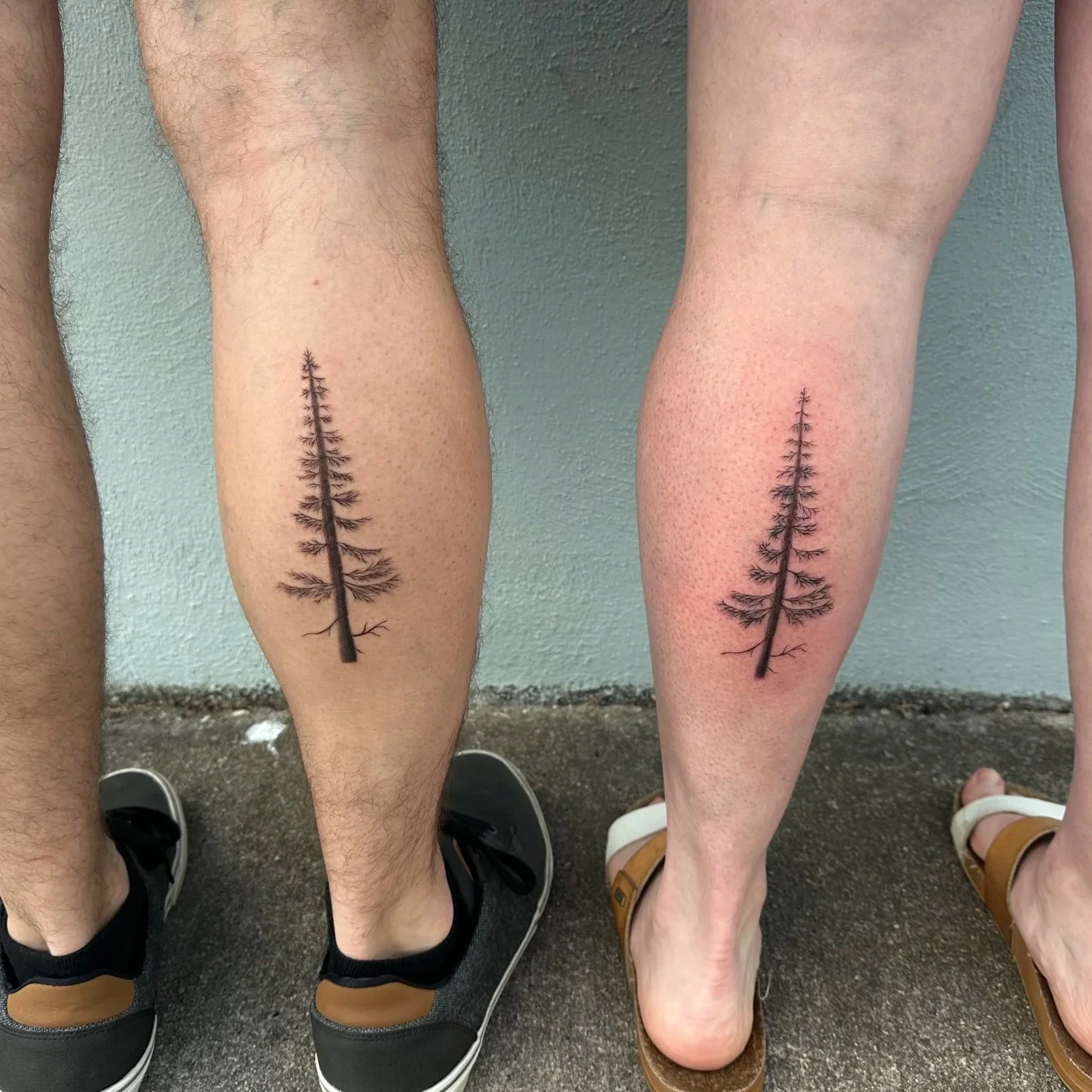 Close-up of two individuals' legs with matching tree branch tattoos, standing on ground with shoes visible. The left person has a hairier leg and black sneakers, while the right person has a less hairy leg, wearing tan sandals.