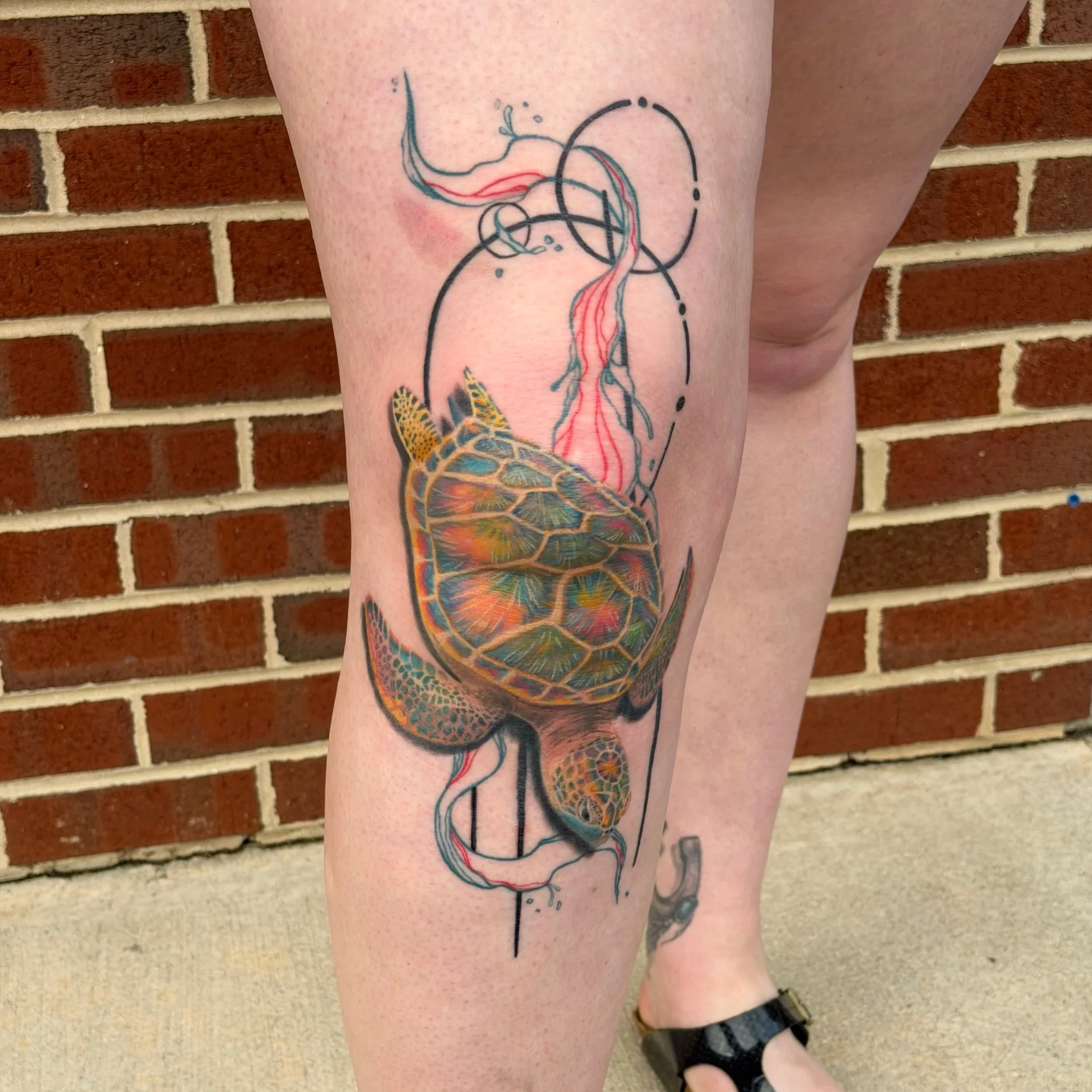 Colorful tattoo of a turtle with a geometric design on a person's thigh, against a brick wall background.