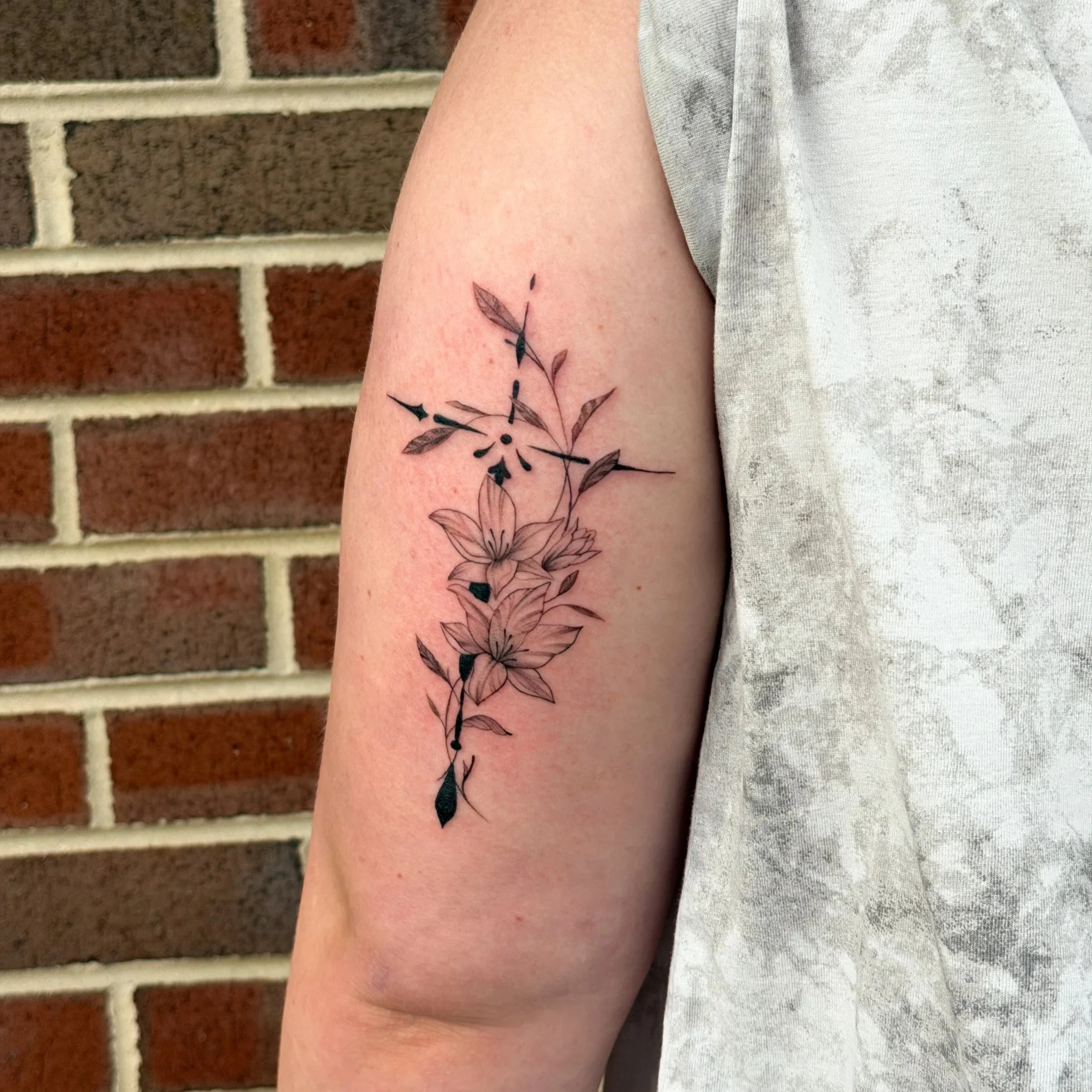 Close-up of a person's arm with a floral tattoo and a gray t-shirt, standing in front of a brick wall.