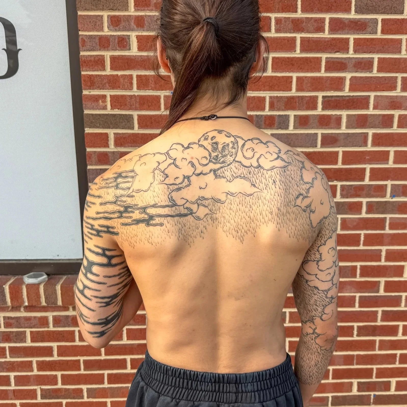 Back of a shirtless person with a detailed tattoo of clouds and a moon on the upper back and arm, standing in front of a brick wall.