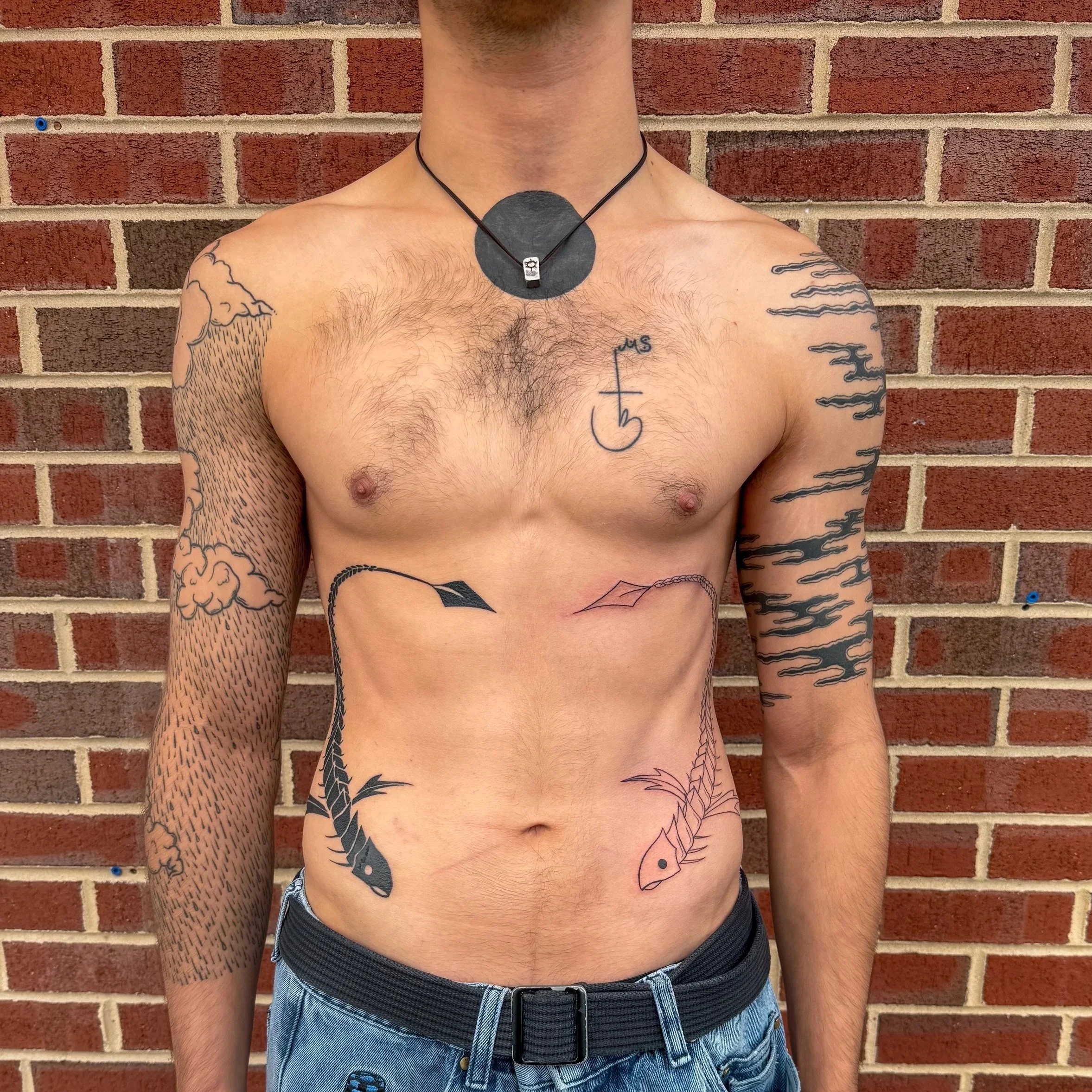 Close-up of a shirtless man standing against a brick wall, wearing a black necklace with a circular pendant and tattoos on his arms and chest.