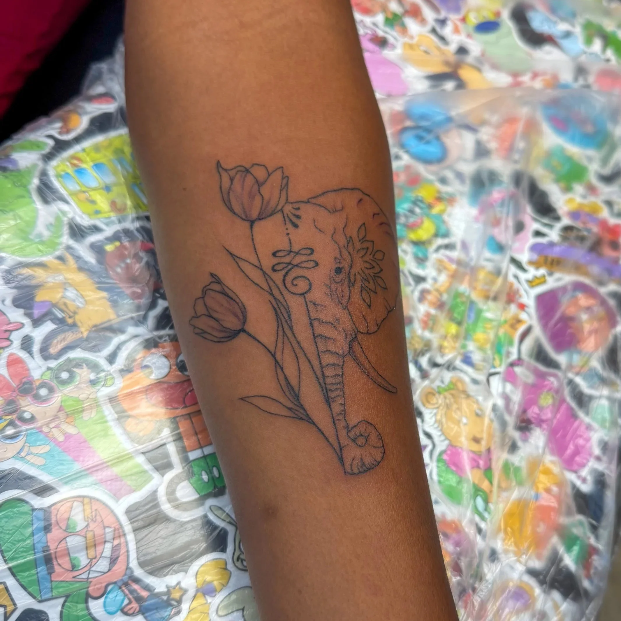 A person’s arm with a tattoo of a tulip flower and some leaves, resting on a surface covered with colorful stickers and decoration.