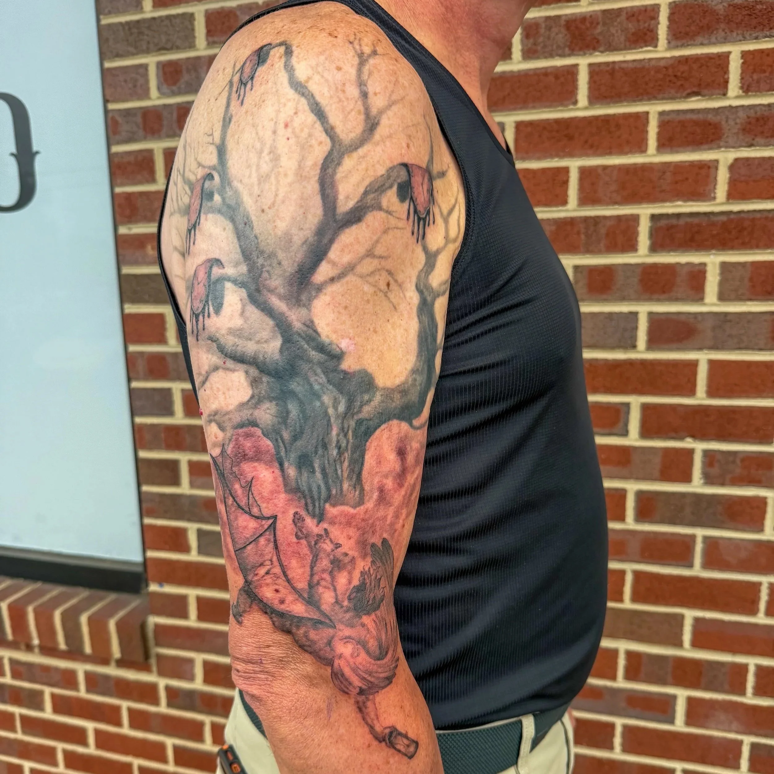 A person with a tattoo sleeve on their upper arm, featuring a tree branch with some hanging objects, against a brick wall background.