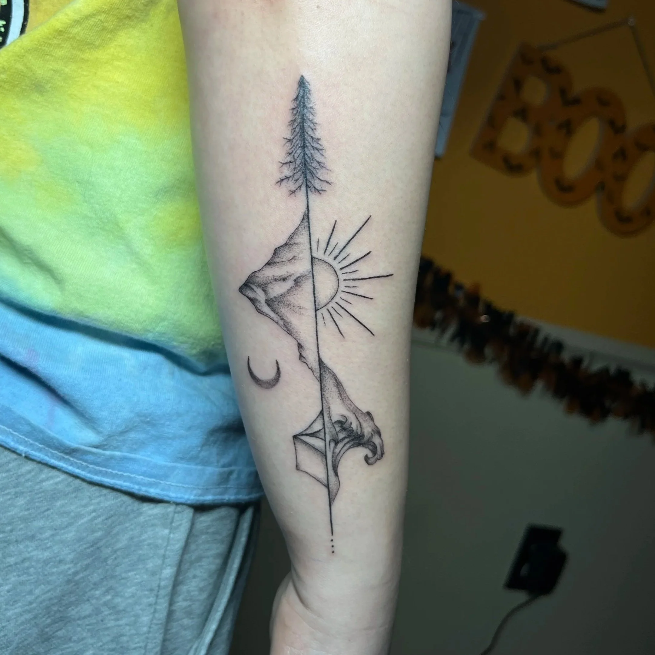 A tattoo of a tree with bare branches is on a person's forearm, along with the sun's rays and a crescent moon.