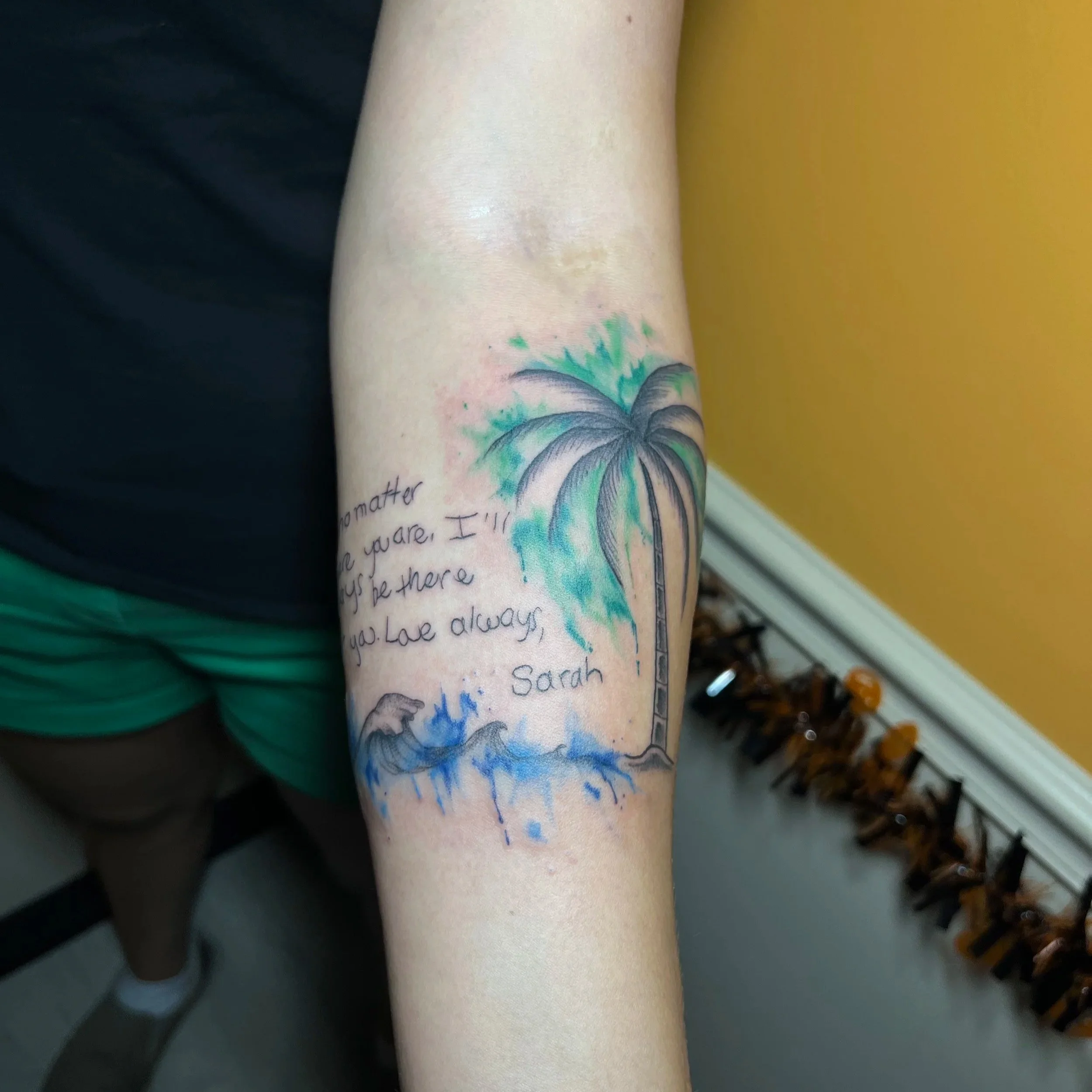 Close-up of a person's arm with a colorful tattoo of a palm tree and ocean waves, and handwritten text on the skin.