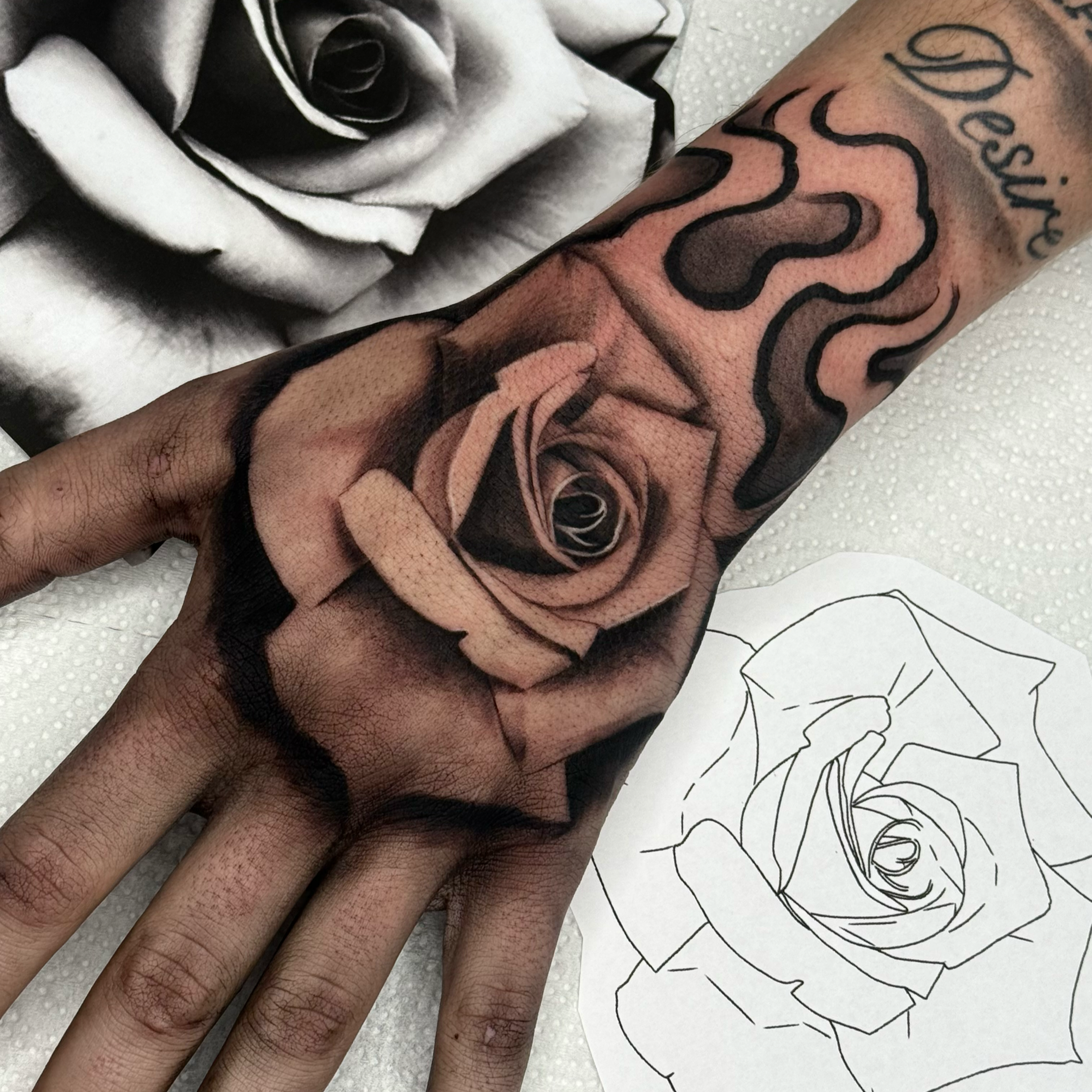 Close-up of a person's arm with a large black and gray tattoo of roses and leaves, with the word 'Beautiful' written in cursive near the top of the tattoo. The tattoo is on the forearm, and the skin appears to be light with some visible hair.