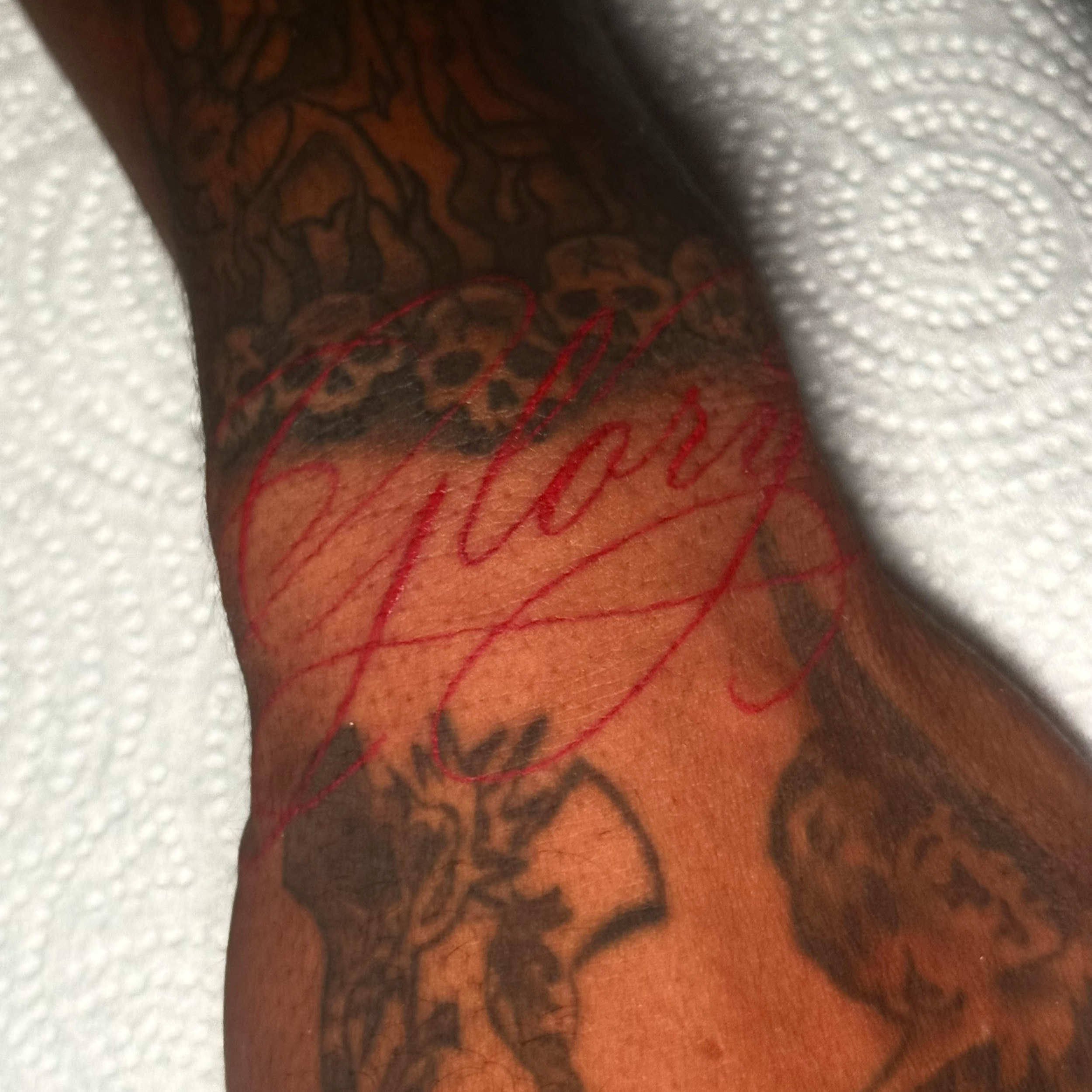 Close-up of a person's arm with a tattoo in black ink, featuring intricate line work and some red text or design on the skin.