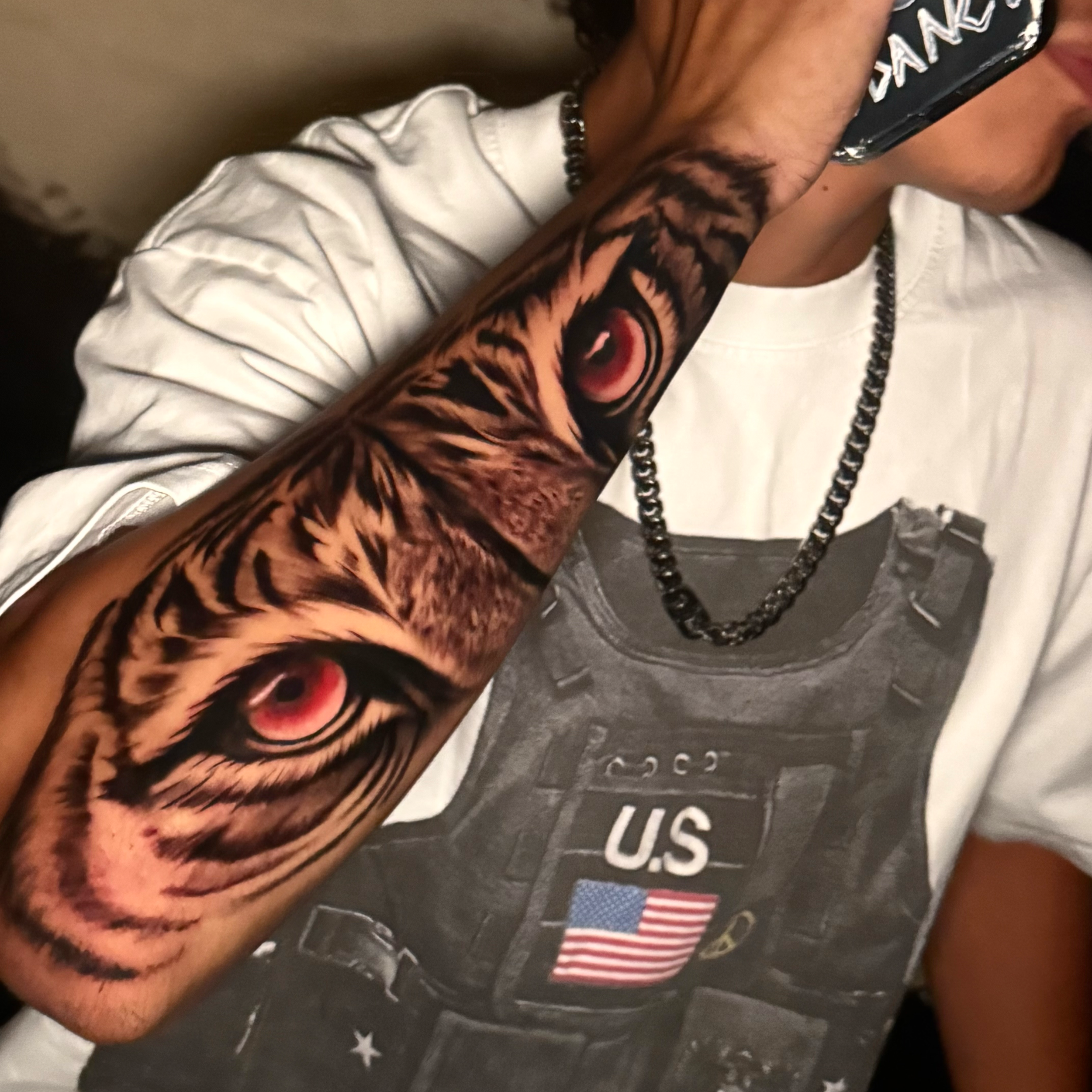 A person's arm with a colorful tiger tattoo, holding a cellphone near their face, wearing a gray shirt with white and red stripes and a beaded necklace with an American flag charm.