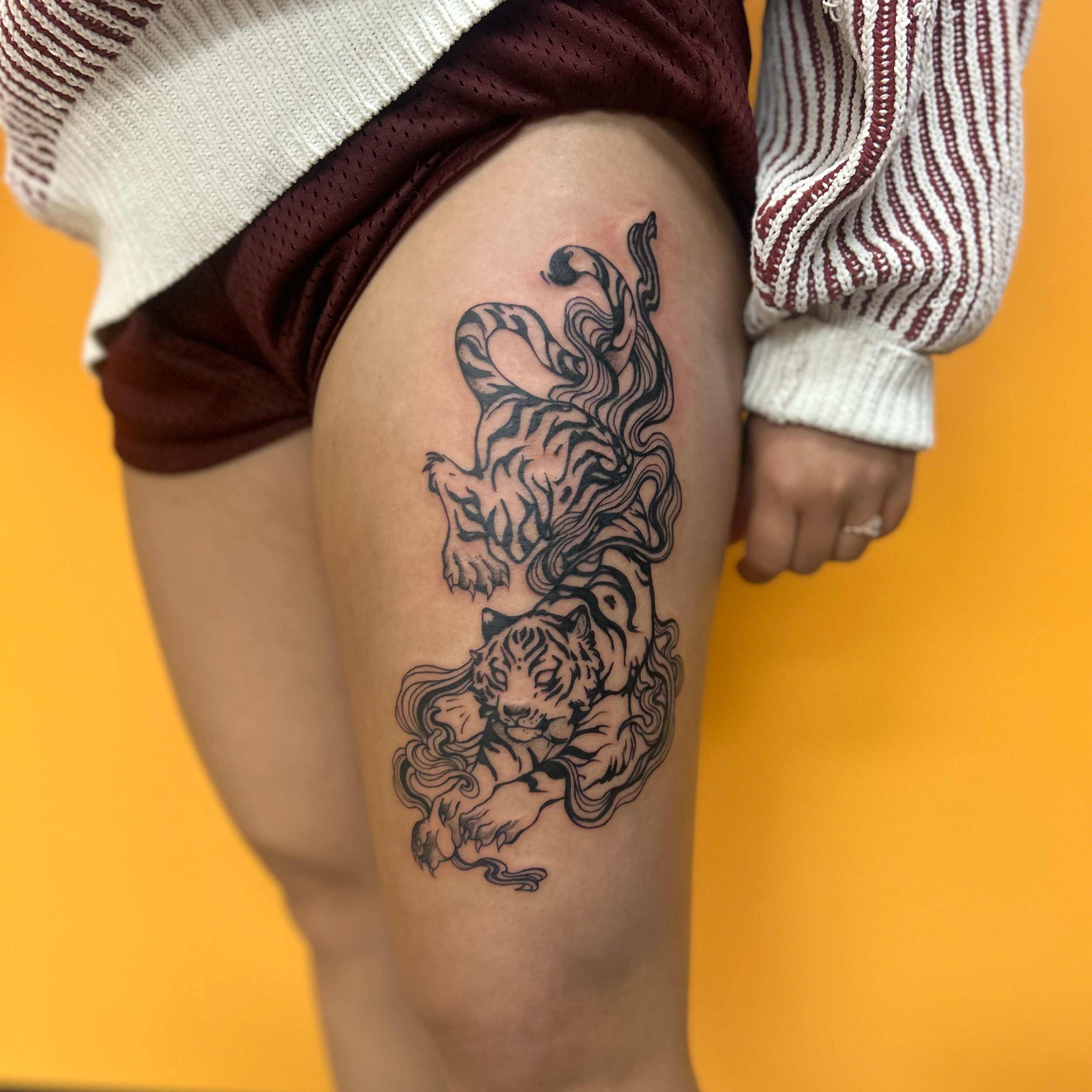 Close-up of a tattoo of a tiger on a person's thigh, with the person wearing burgundy shorts and a striped jacket, against a yellow background.