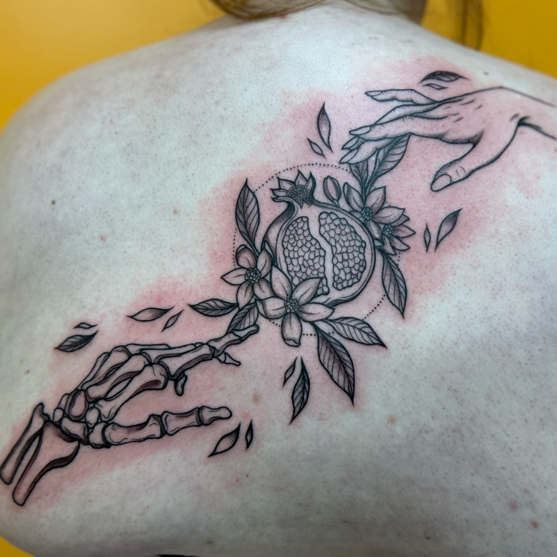 A black outline of a pomegranate surrounded by floral elements and a hand reaching towards it, tattooed on a person's shoulder.