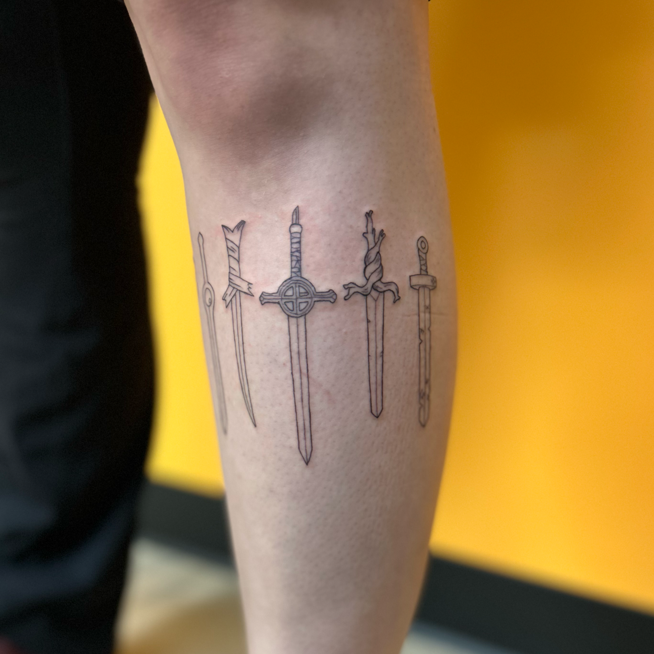 A person's forearm with a tattoo of multiple small swords or knives, each with a different design, arranged in a row with a yellow background.