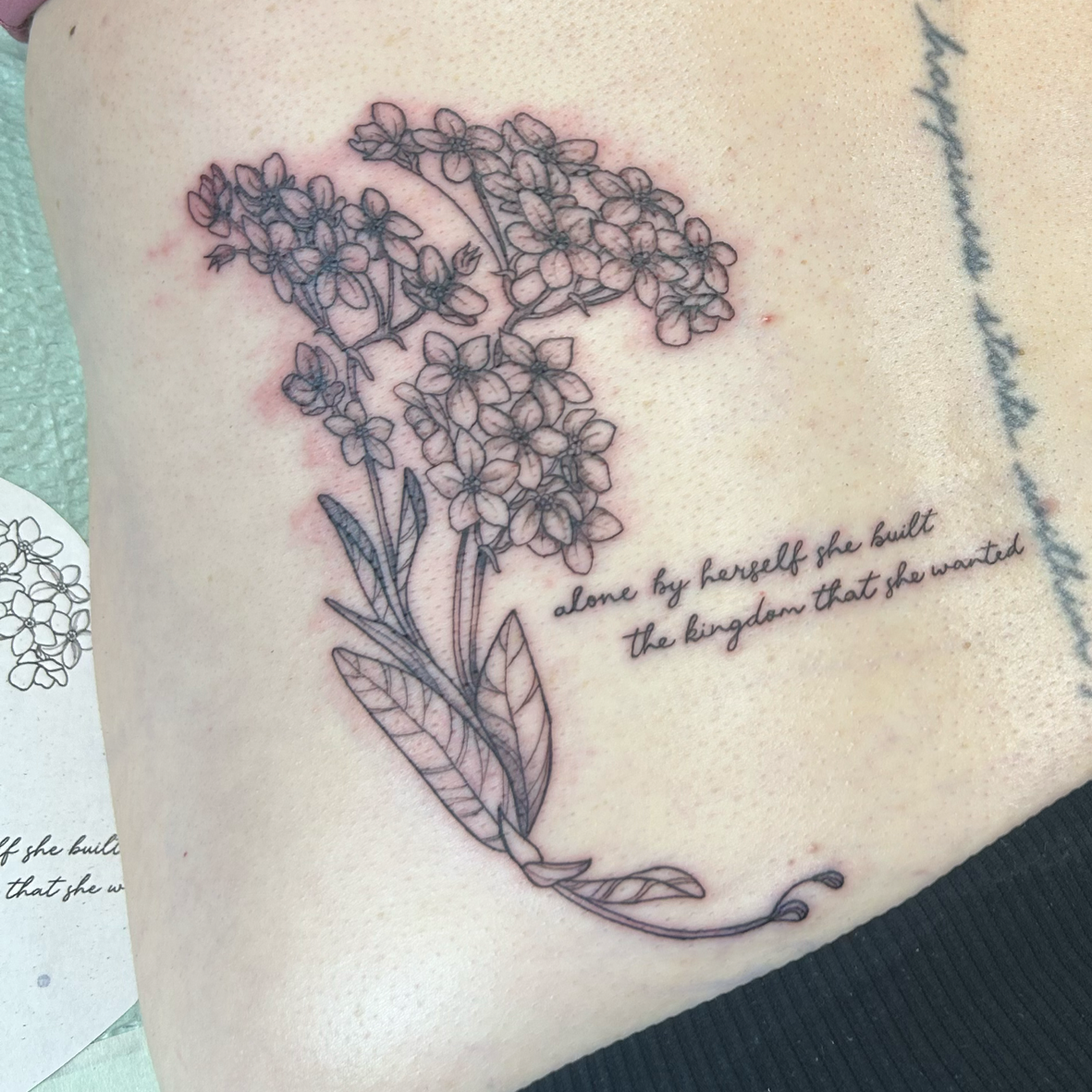 A black line tattoo of a flowering plant, with a quote that reads "alone by herself she built the kingdom that she wanted."