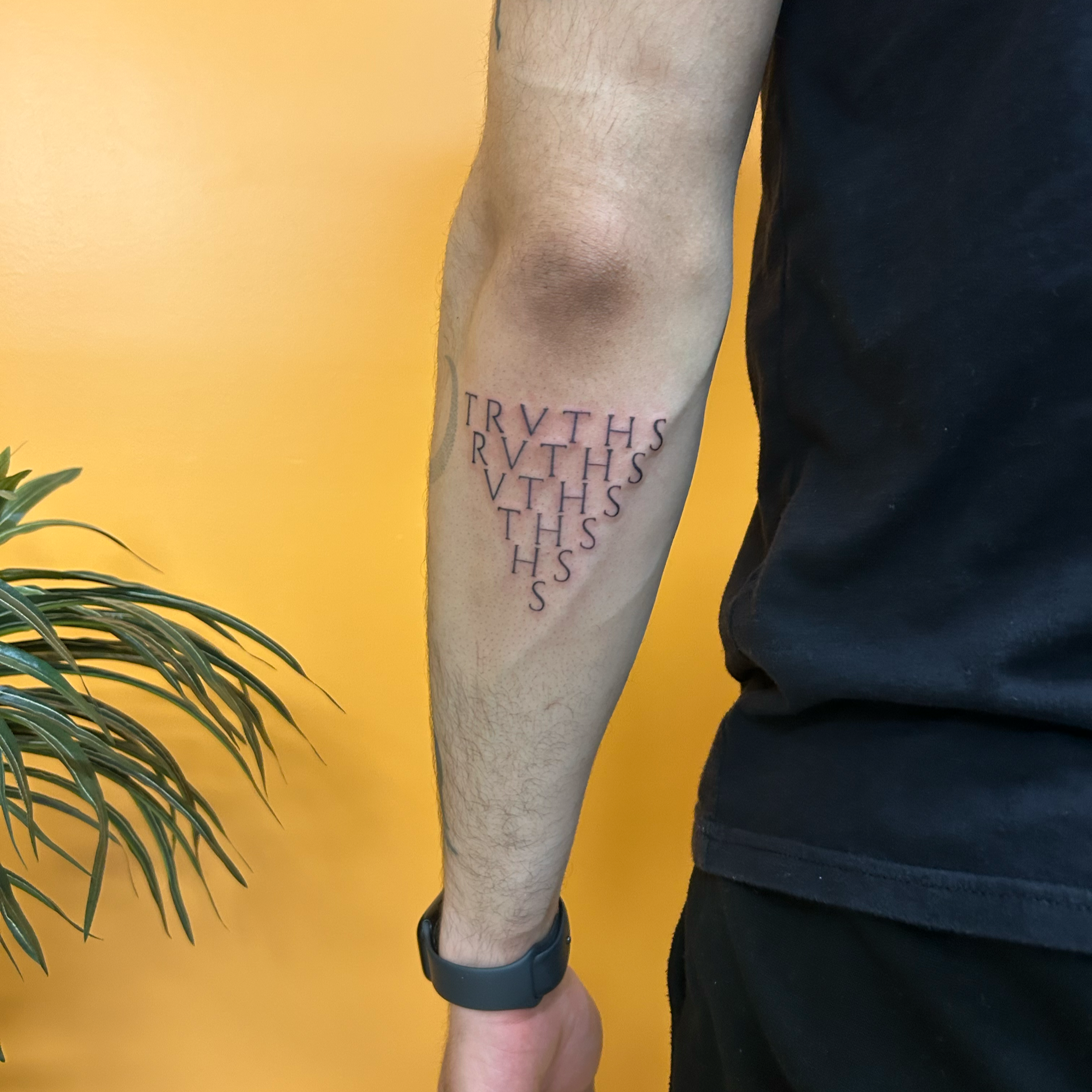 Close-up of a person's arm with a tattoo that says "TRVTHS" repeated multiple times in black ink, against a yellow background. Part of a black shirt and a black watch are visible, with some greenery in the lower left corner.