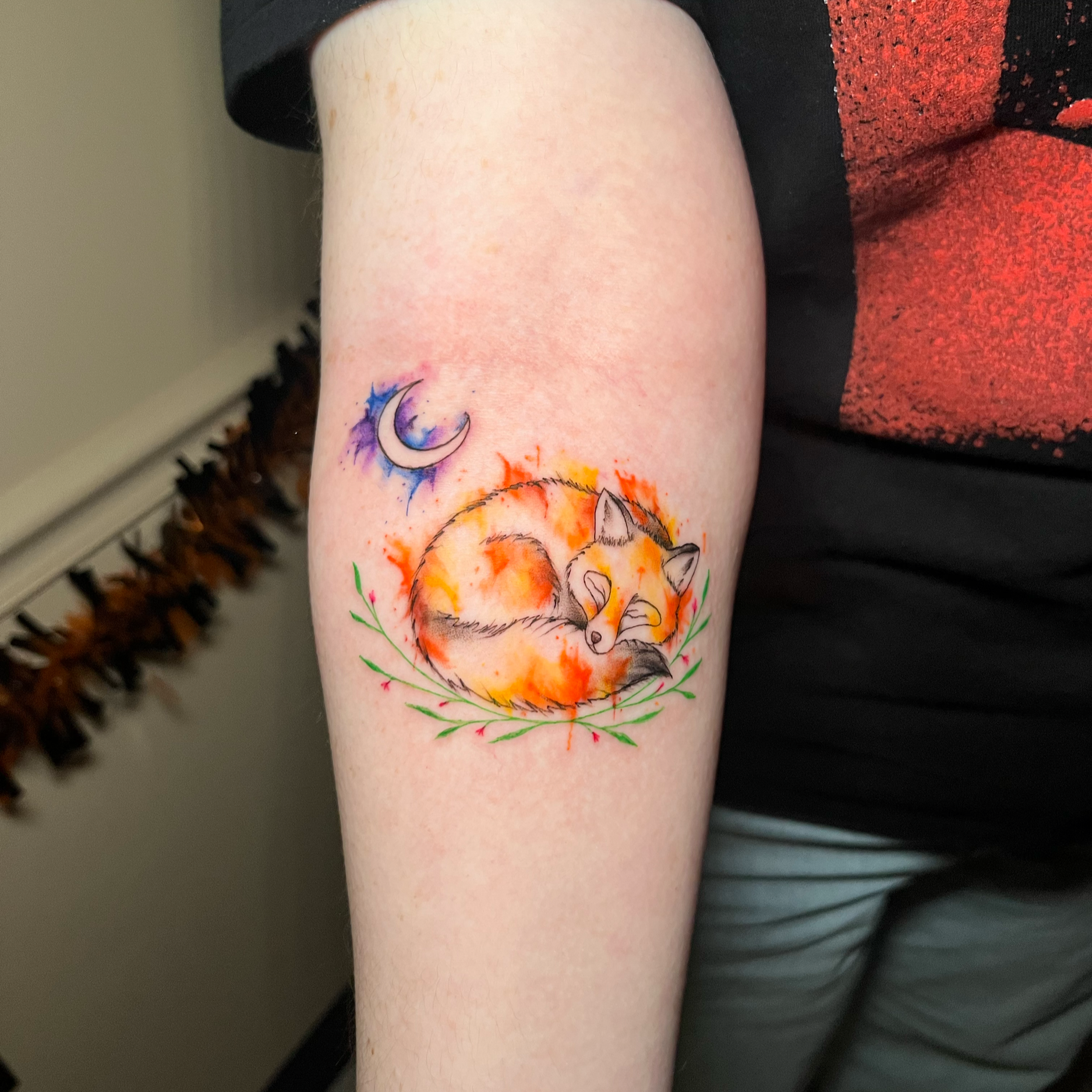 Tattoo of a crescent moon with watercolor-like purple and blue splashes on the skin above an orange flame-colored circle with a fox curled within, surrounded by green branches on a person's arm.