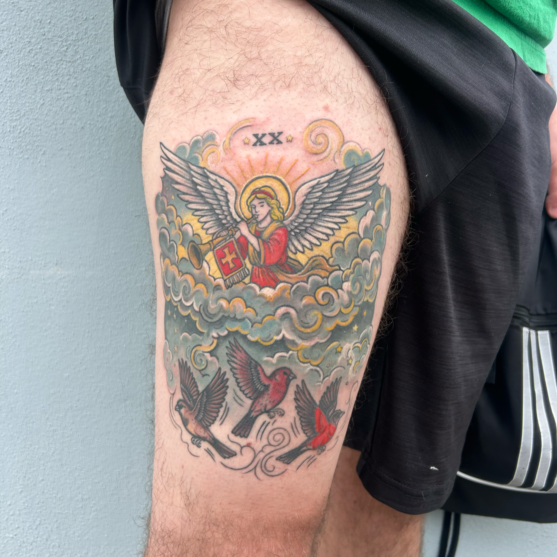 Tattoo of an angel with wings, holding a shield with a cross, surrounded by clouds and the text 'XX' at the top, on a person's upper arm.
