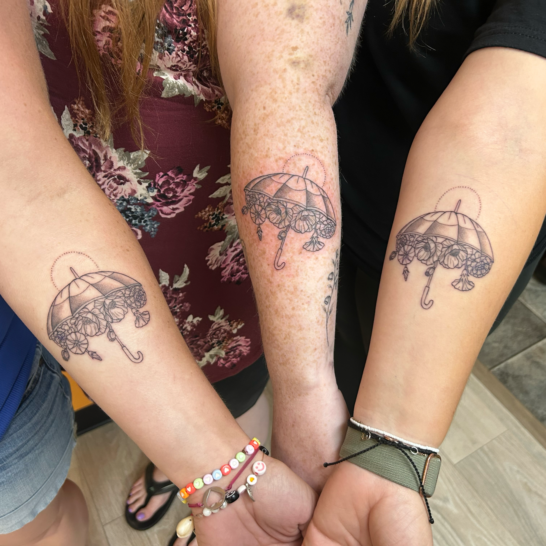 Close-up of four people's arms with matching umbrella tattoos. The people are wearing various bracelets and casual clothing, standing close together indoors.