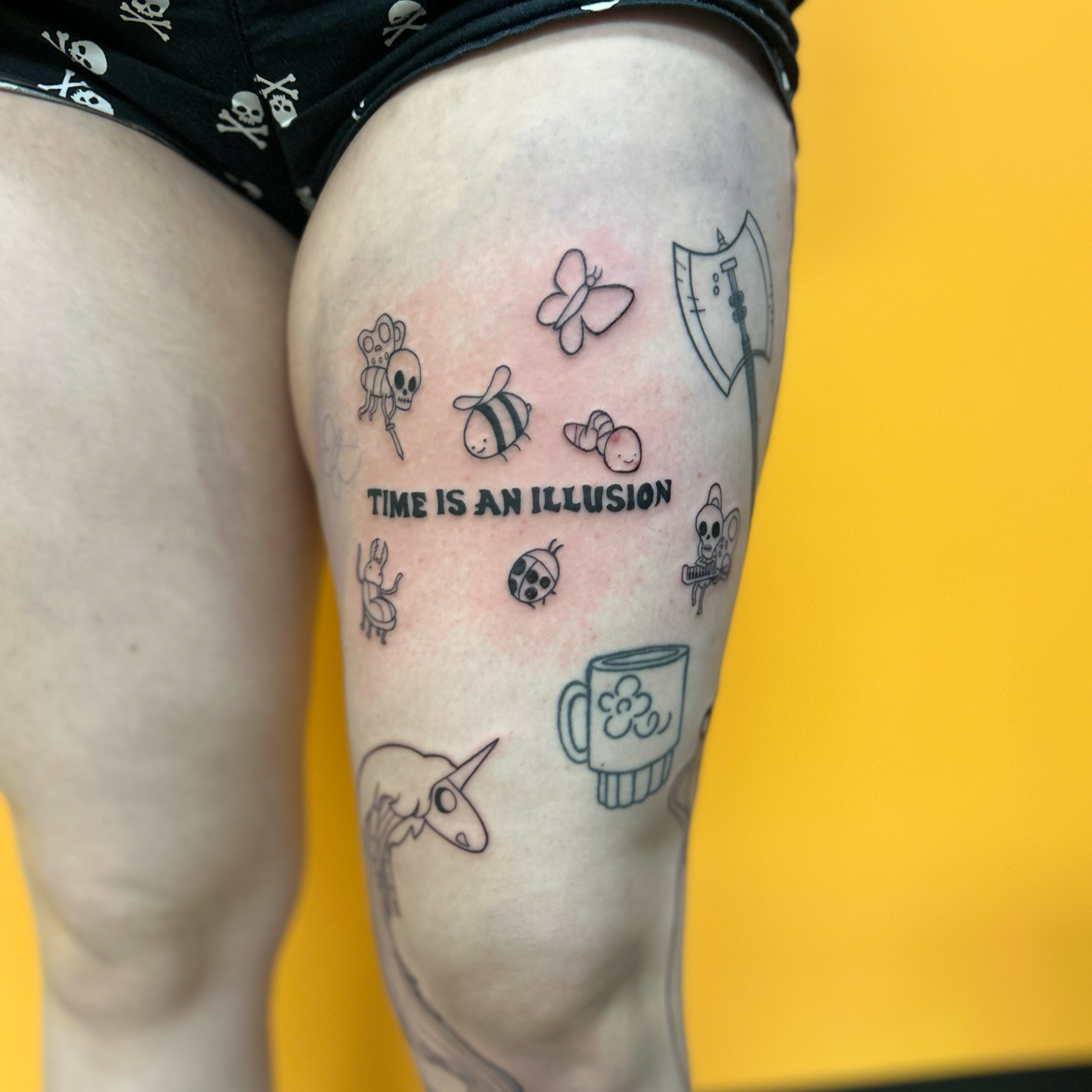 Close-up of a person's thigh with various small, black outline tattoos, including a skull, a butterfly, a bee, and a mug of beer. The person is wearing black shorts with skull and crossbones patterns, and the background is yellow.