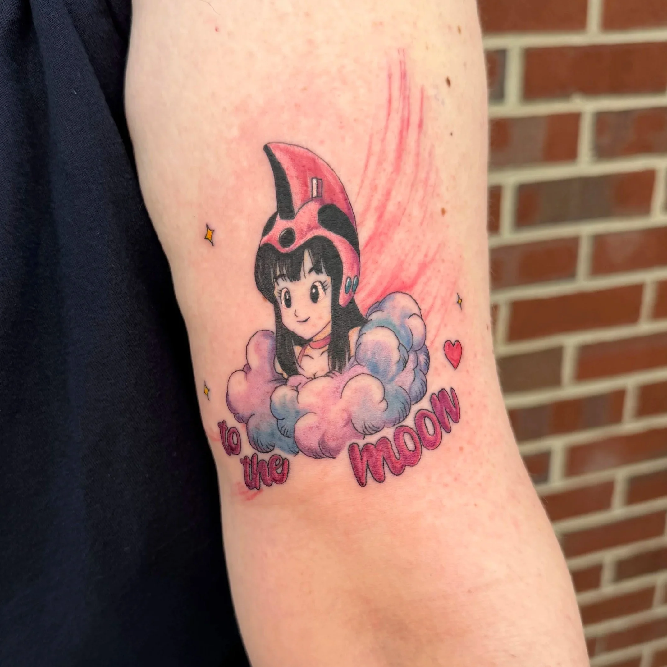 Tattoo on person's arm featuring a cartoon girl with dark hair, pink helmet, and clouds, surrounded by small yellow stars and red hearts, with pink text.
