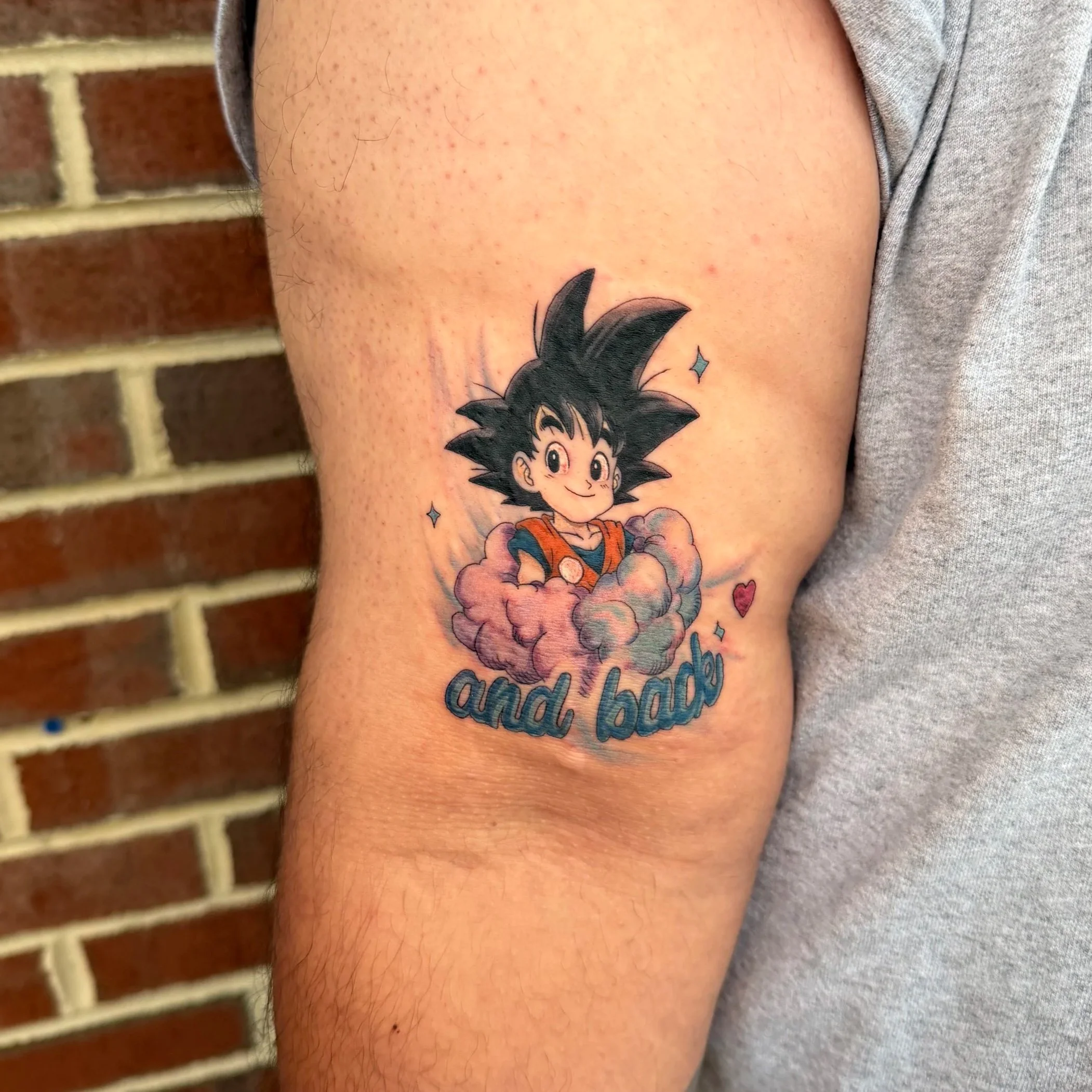 Close-up of a person's arm with a tattoo of Goku from Dragon Ball Z surrounded by pink clouds and surrounded by colorful stars, with a brick wall in the background.