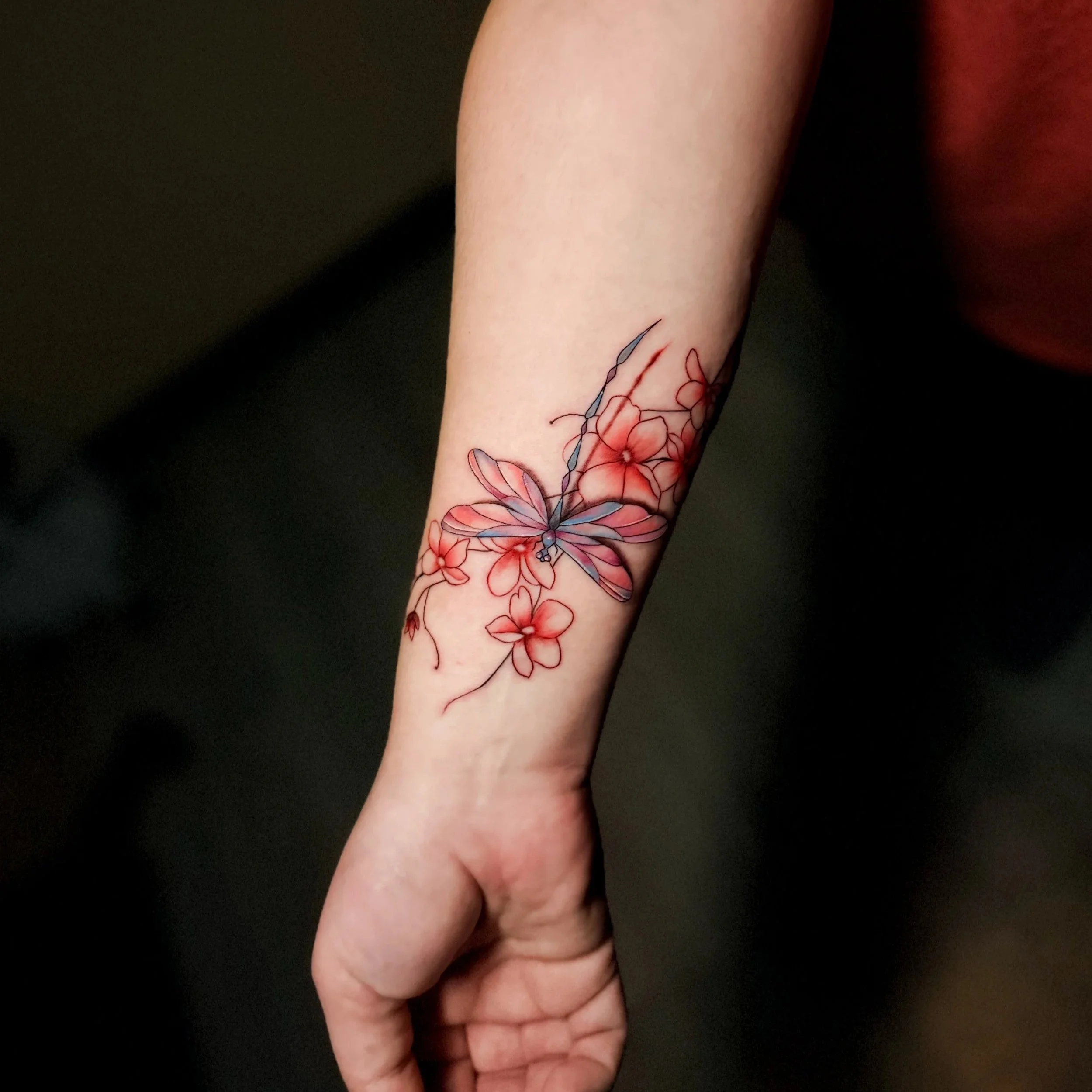 Close-up of a forearm with a floral tattoo featuring red and pink flowers and blue leaves, against a black background. dragonfly and cherry blossoms
