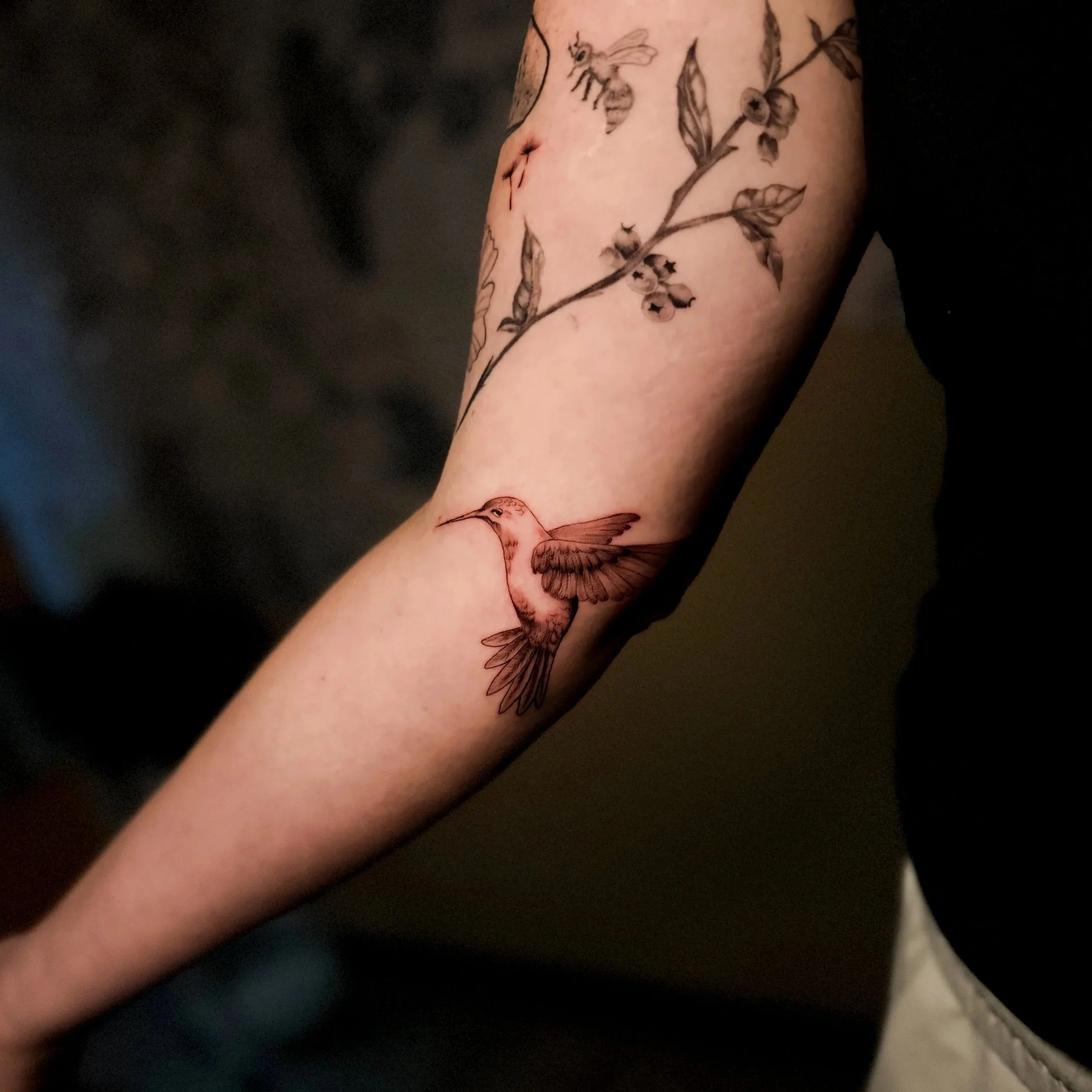 A tattoo of a hummingbird on a person's forearm, with a floral branch and bee in black ink and fine line style.