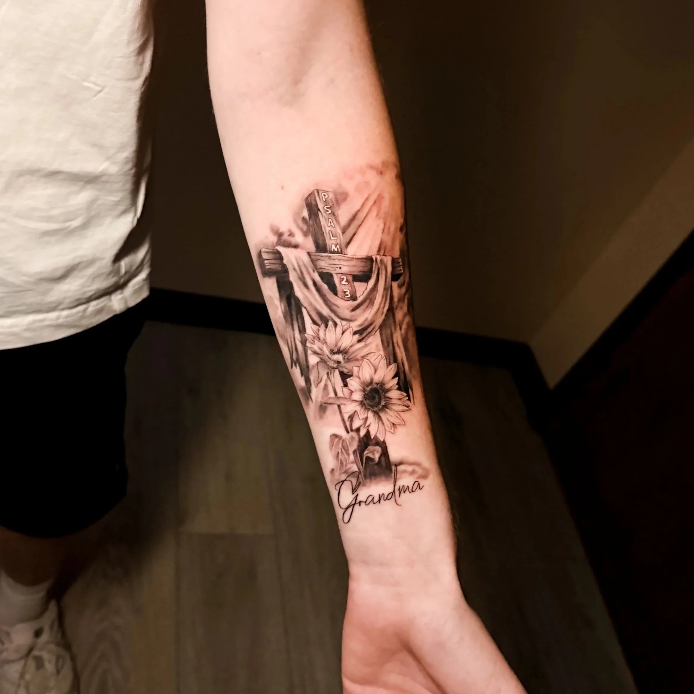 A tattoo of a wooden cross with the words PSALM 23 on it, surrounded by flowers on the person's forearm.