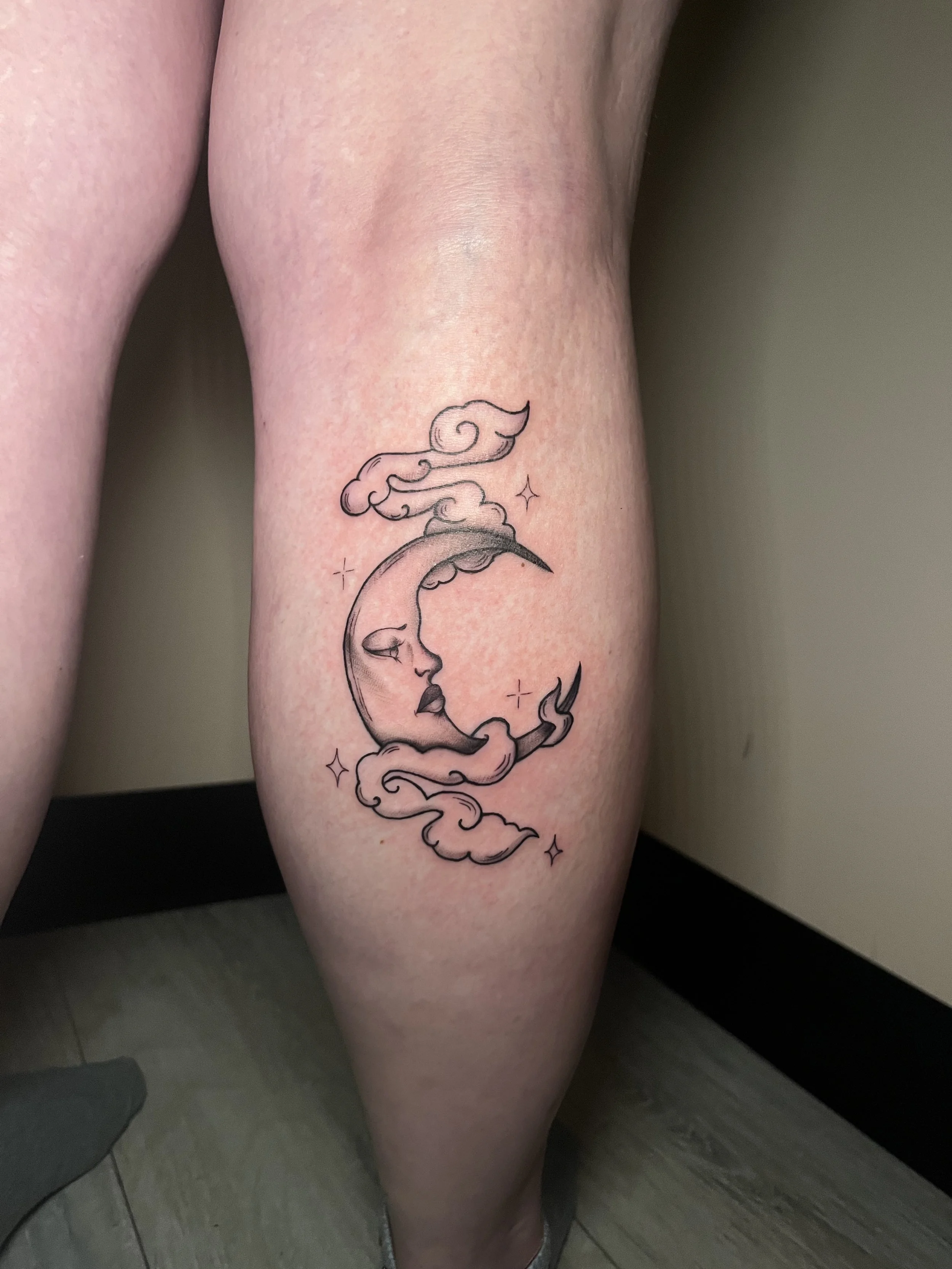A tattoo of a crescent moon with a face, surrounded by clouds and small stars, on a person's thigh.
