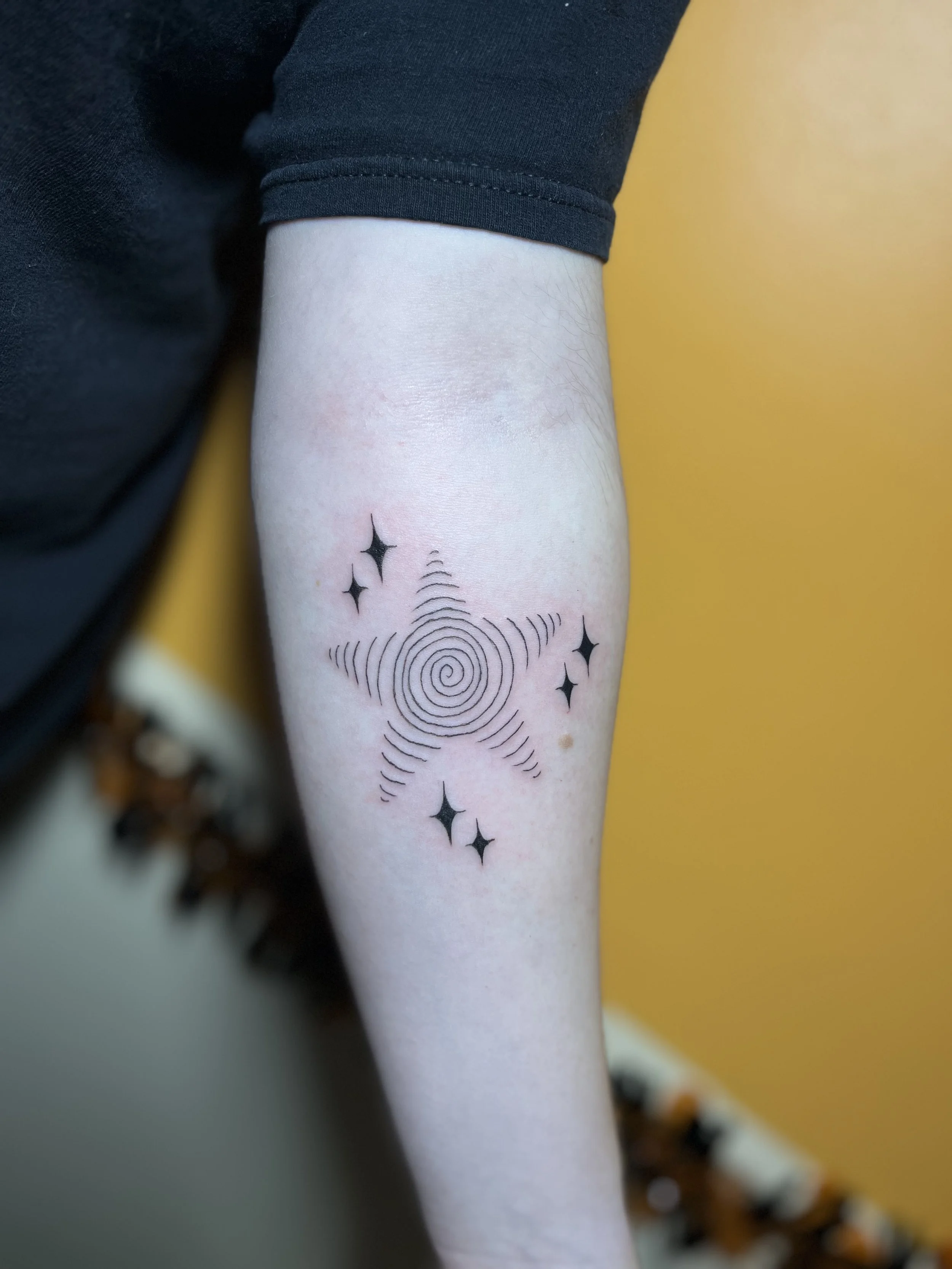 Tattoo of a spiral with radiating lines and four small stars on a person's forearm against a yellow background.