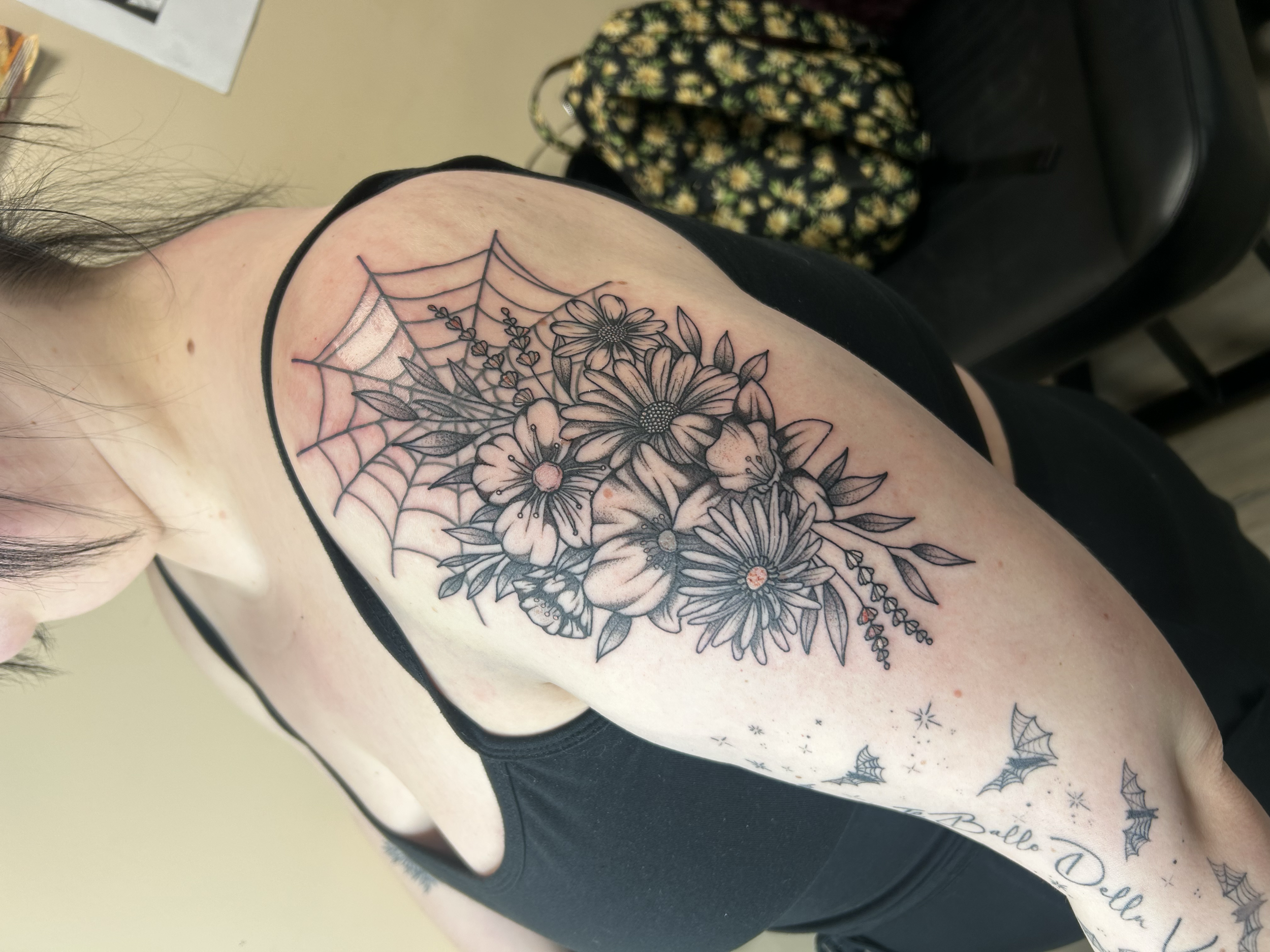 Black and gray floral tattoo with spider web and small designs on a person's shoulder and upper arm.