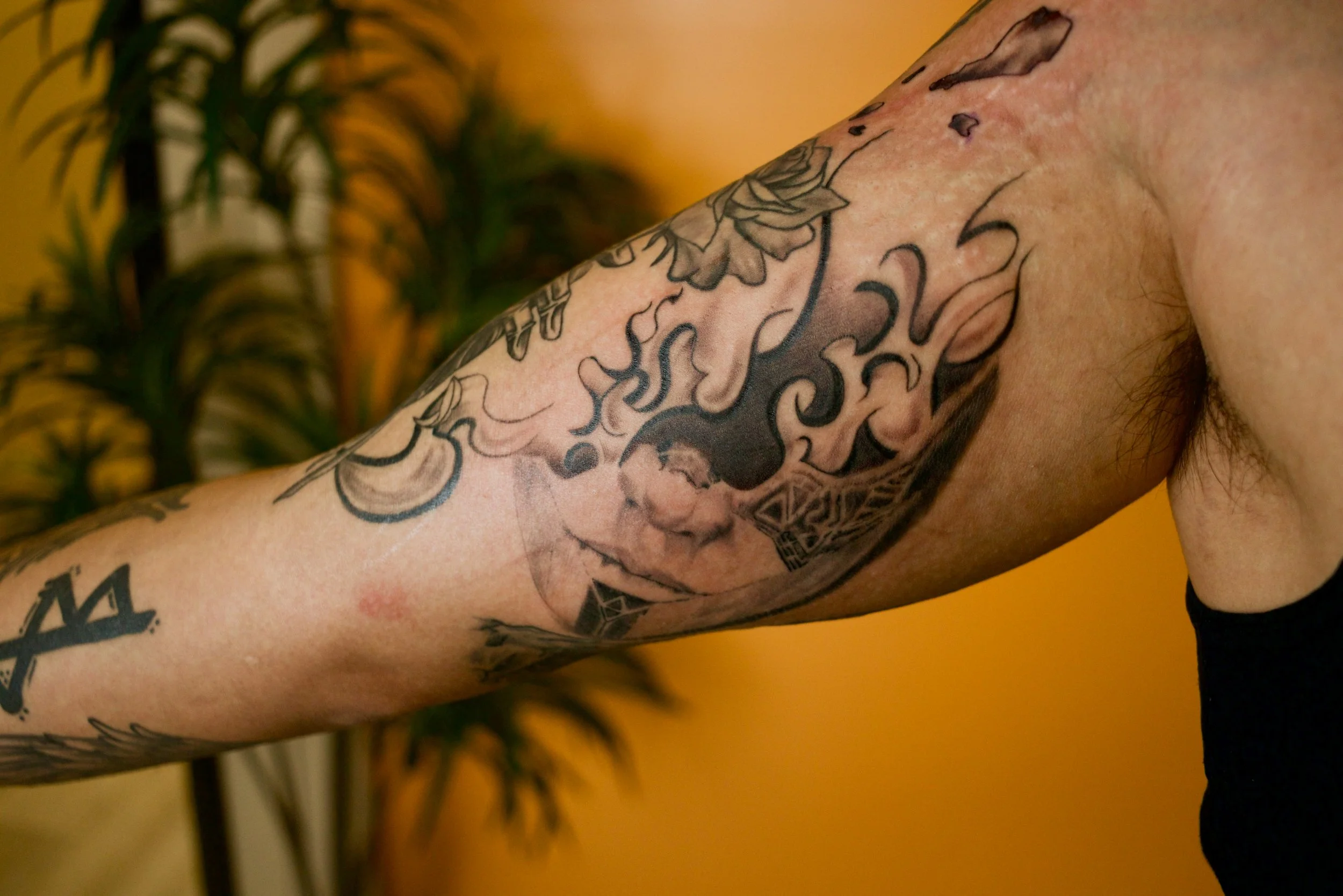 Close-up of a person's upper arm and shoulder with multiple tattoos, including a large face, abstract designs, and symbols. The background features a yellow wall and a blurred green plant.